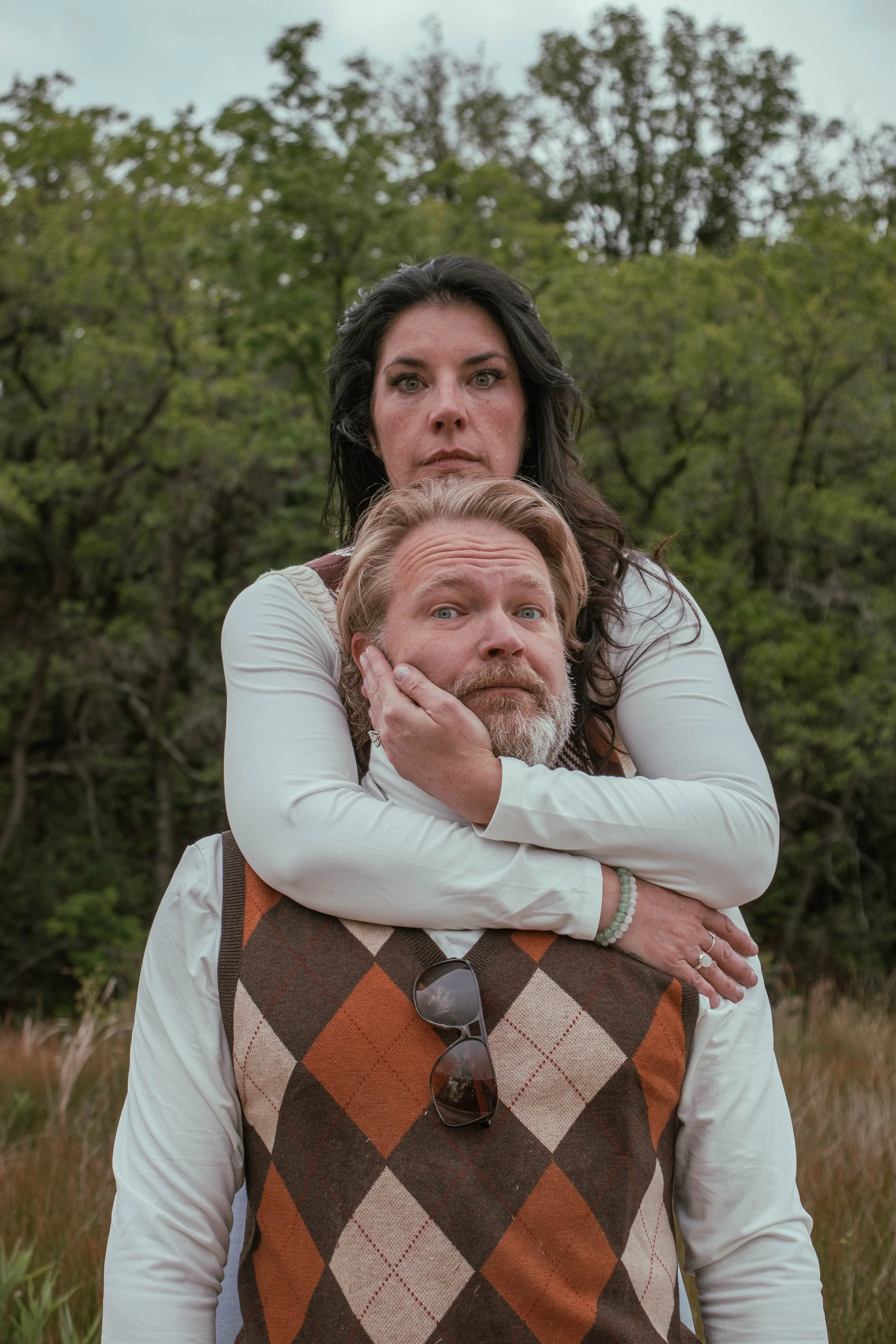 A woman with dark hair and a man with a beard and mustache in a close, affectionate pose outdoors surrounded by trees and greenery.