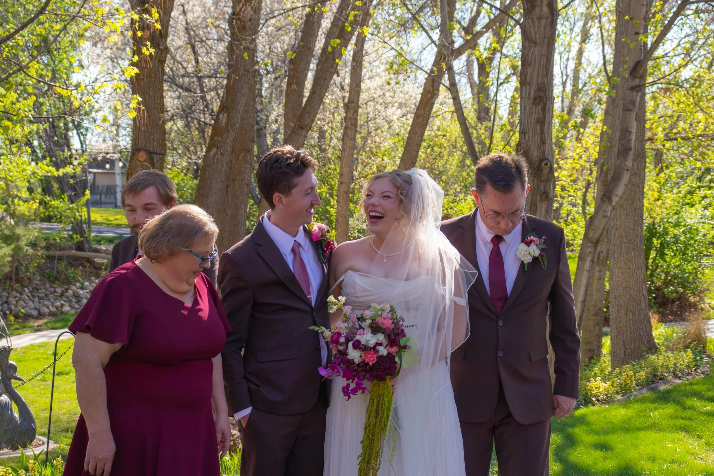 A bride and groom standing outdoors with three older adults, likely family members, during a wedding ceremony. The bride wears a white dress and veil, holding a bouquet of pink and purple flowers. The groom is in a dark suit with a pink tie. The scen