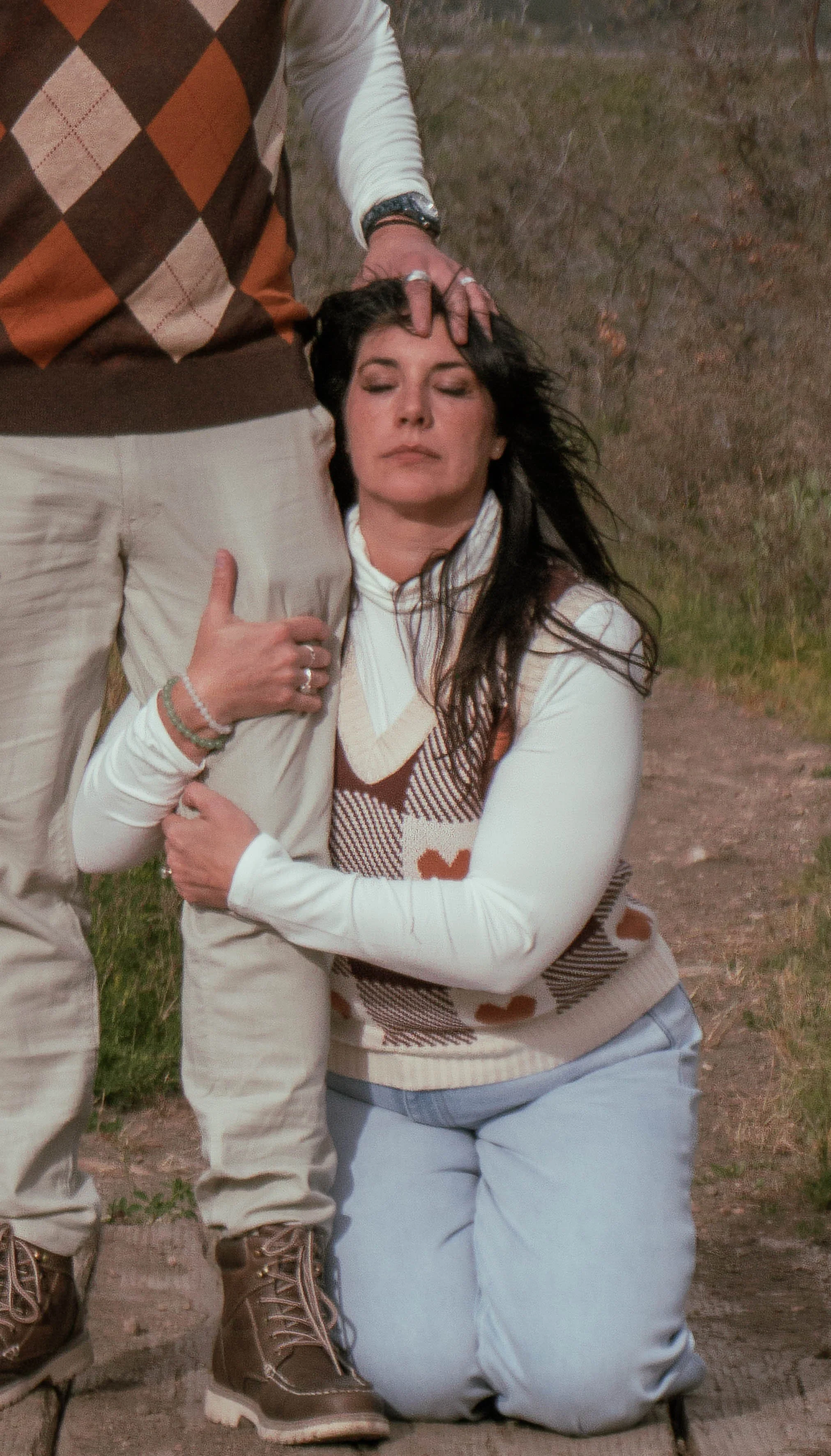 A woman with long dark hair, wearing a knitted vest and light blue jeans, kneeling on the ground outdoors with eyes closed, holding onto a man's leg, who is standing beside her. The man is wearing light-colored pants, a vest with a checkered pattern,