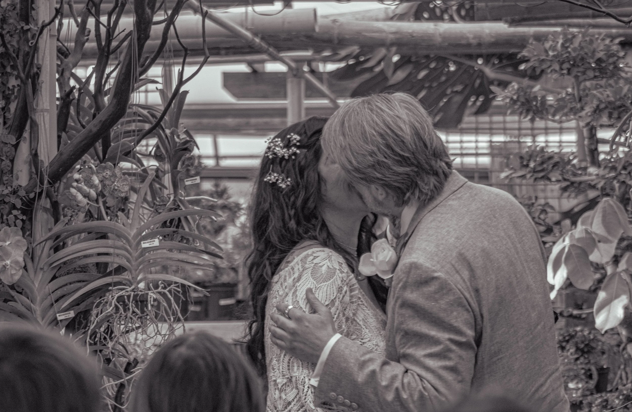 An elderly couple sharing a kiss in a greenhouse with plants and flowers around them.