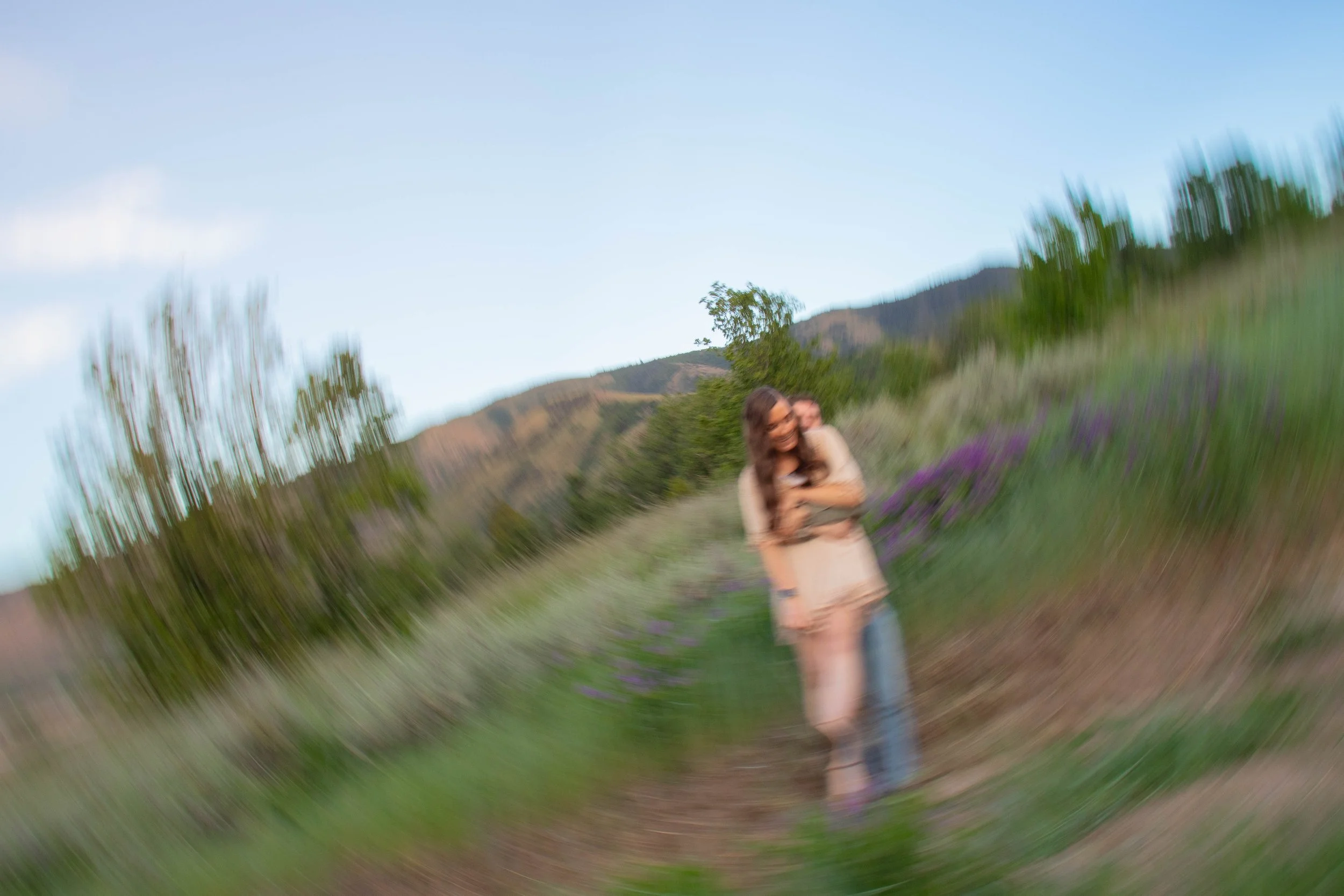 A woman and a child smiling and hugging outdoors in a field with purple flowers, with mountains and trees in the background and a clear blue sky.