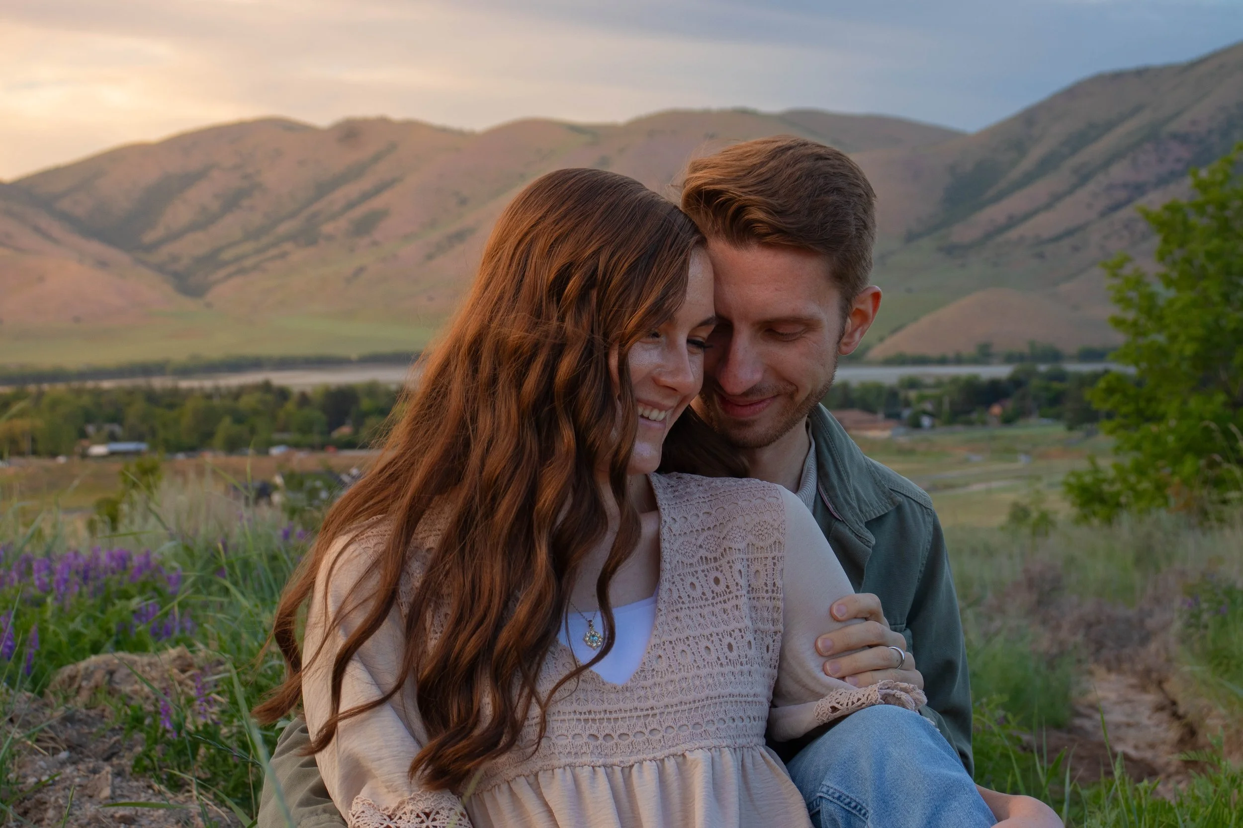 A young couple is sitting close together outdoors, smiling and showing affection with their foreheads touching. They are surrounded by a scenic landscape of green hills, a body of water, and a partly cloudy sky.