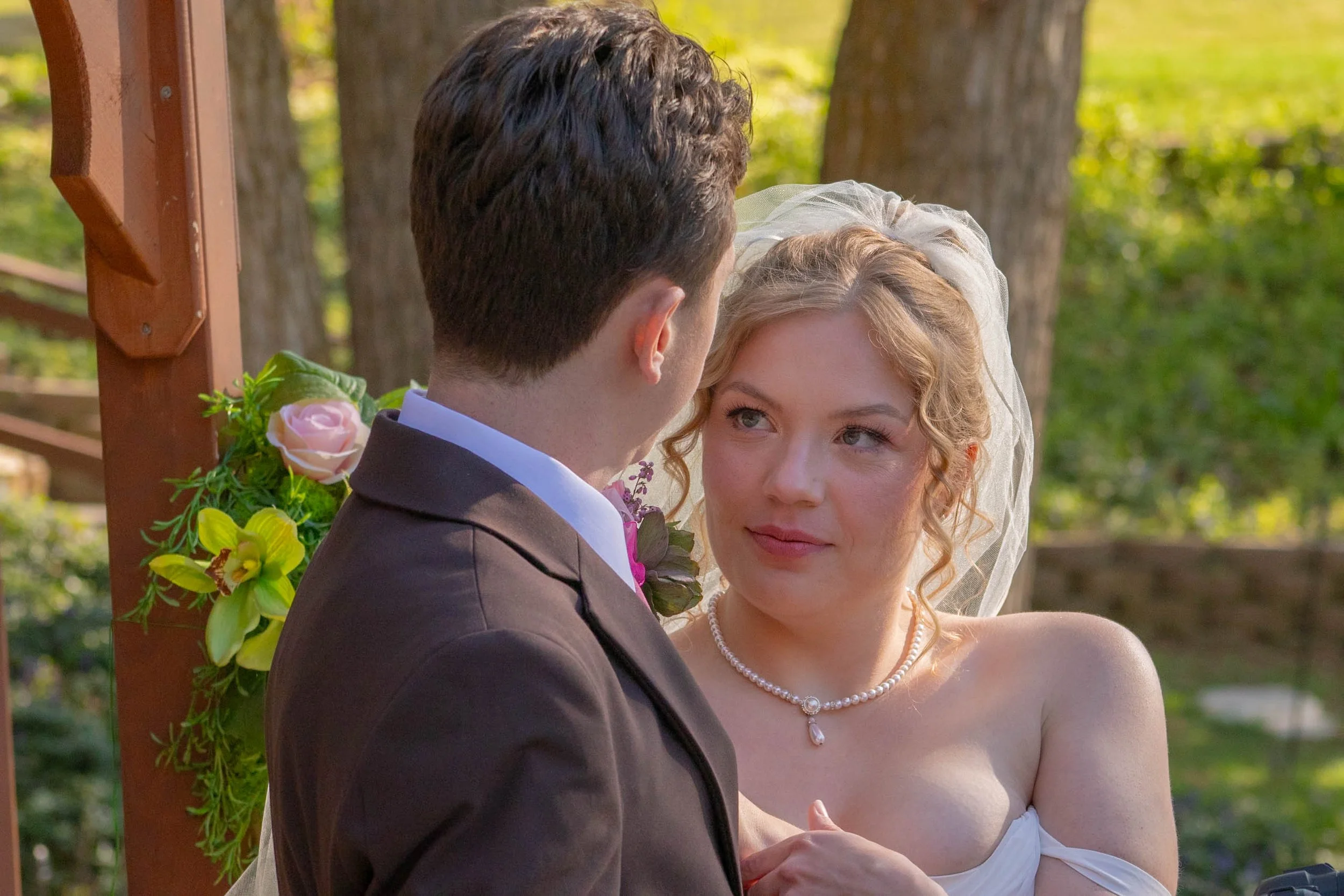 A bride and groom are standing outside in front of a wooden arch decorated with yellow and pink flowers. The groom has dark, curly hair and is wearing a black suit with a white shirt. The bride has blonde hair styled in loose curls, wears a strapless