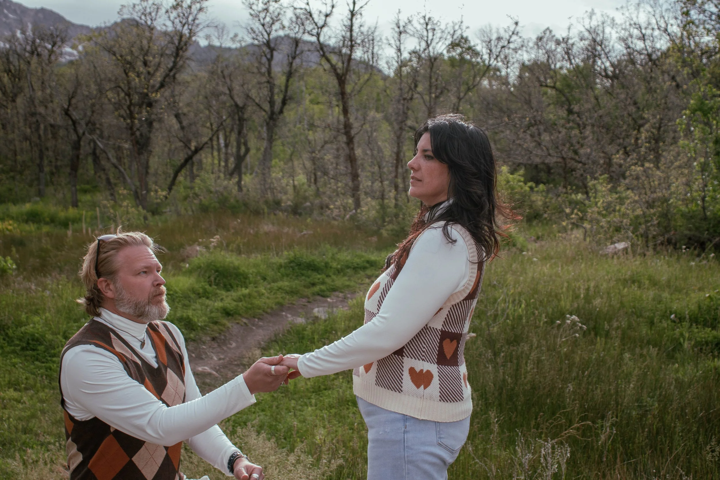 A man on bent knee holding a woman's hands, she stands facing him outdoors in a green, wooded area.
