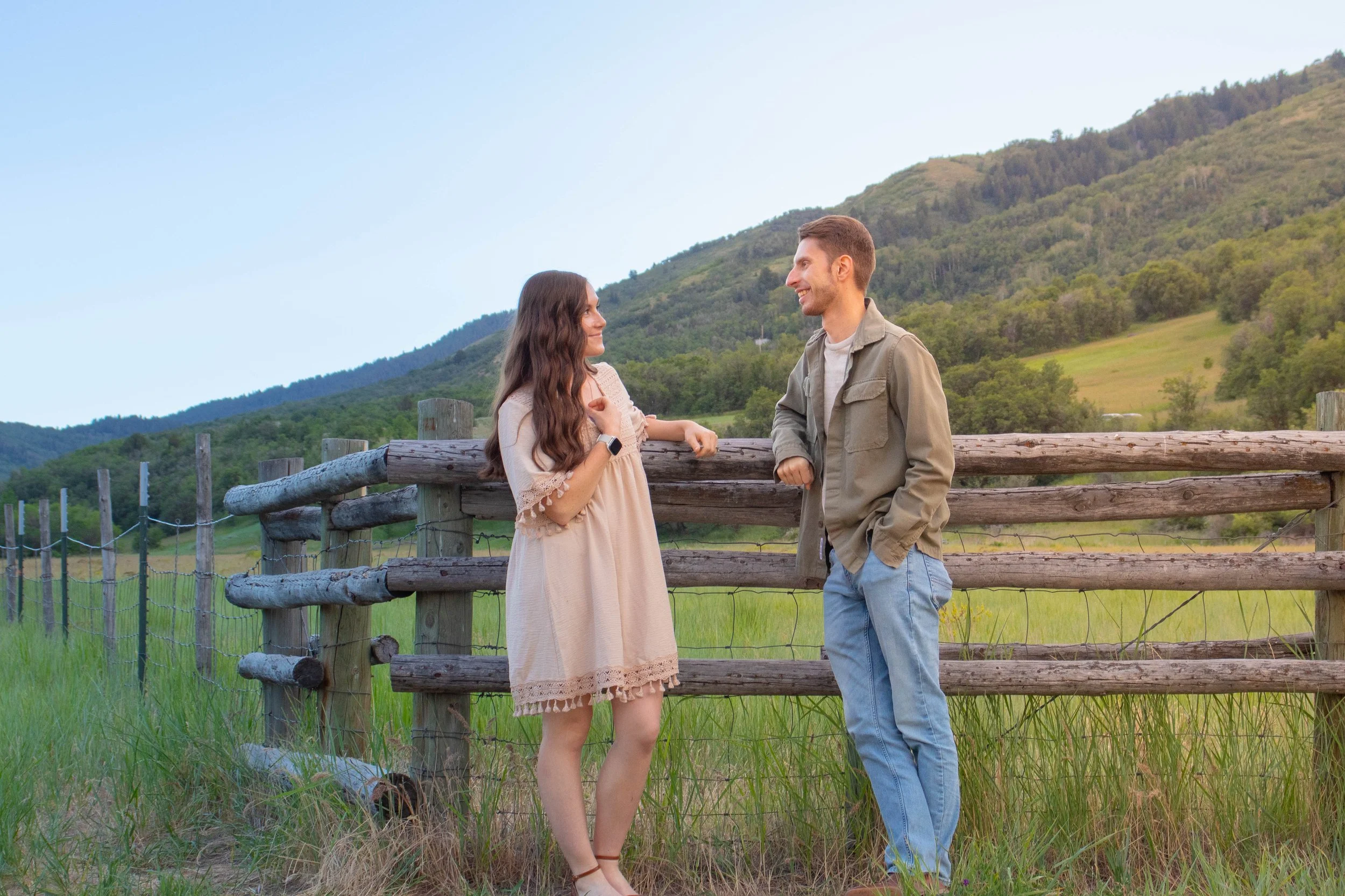 A man and woman stand facing each other by a wooden fence in a rural setting with mountains and trees in the background. They are smiling at each other.