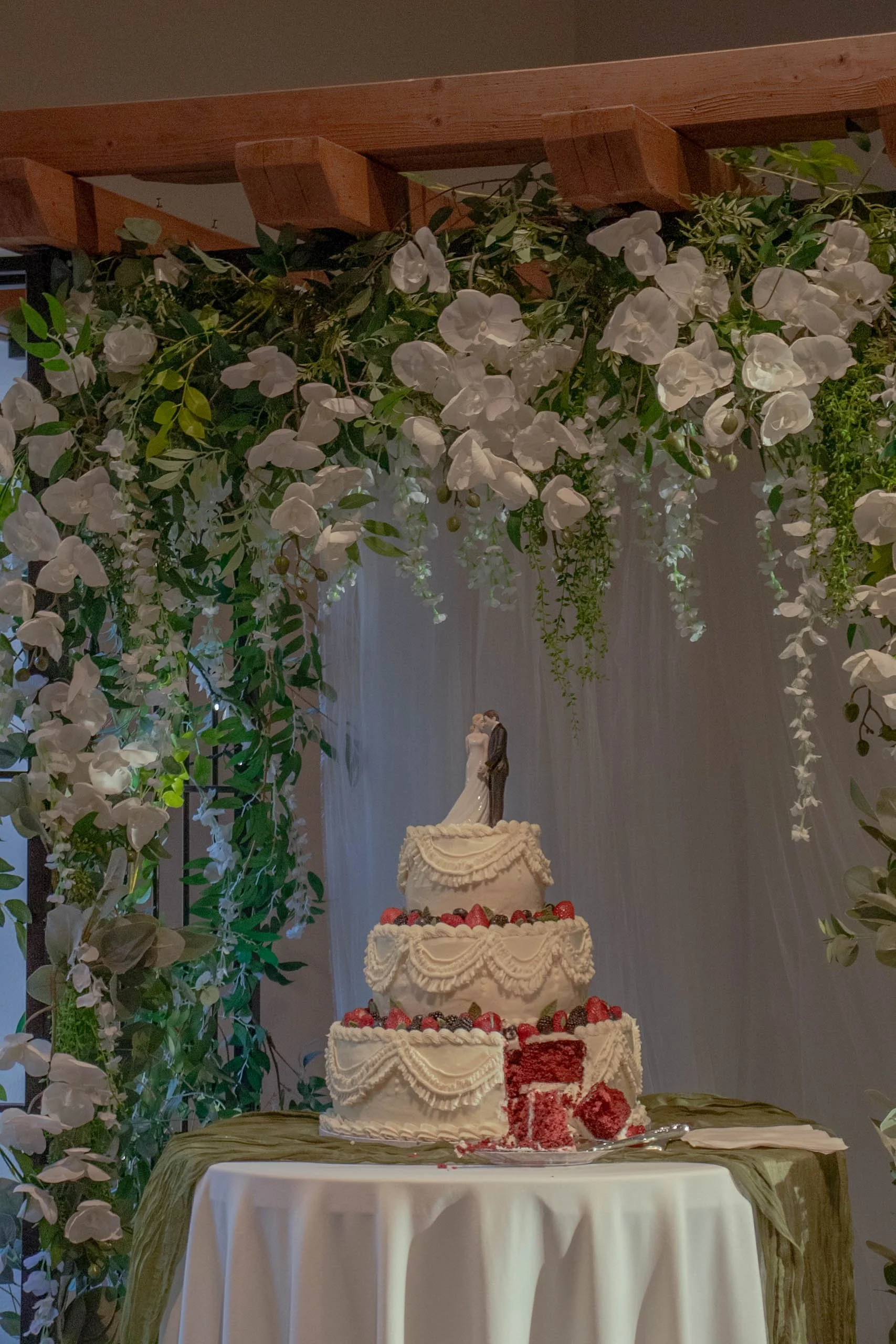 A multi-tiered wedding cake decorated with white icing, strawberries, and other berries, placed on a table with a green cloth. Overhead, there is a floral arrangement with white flowers and green foliage hanging from a wooden loft, with a statue of a bride and groom visible in the background.