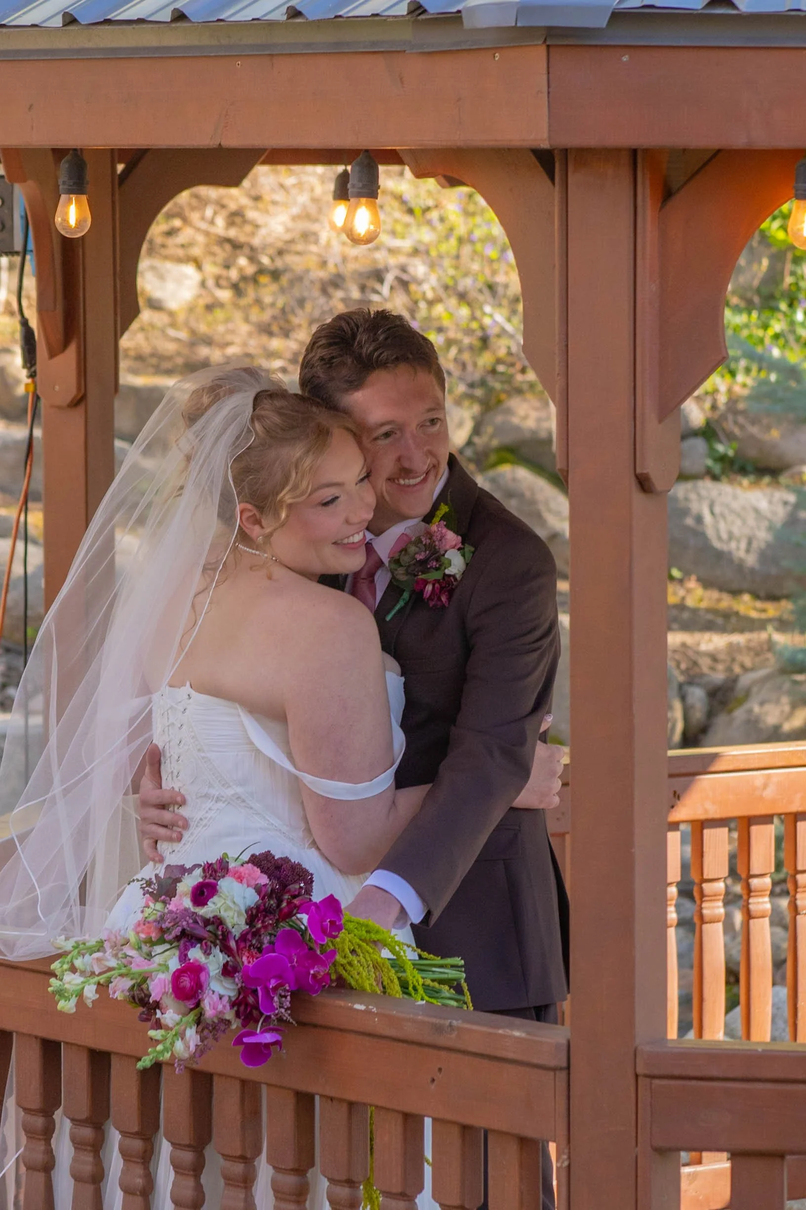 A newlywed couple in formal wedding attire sharing a hug and smiling on a wooden balcony decorated for a wedding, with a bouquet of vibrant pink and purple flowers.