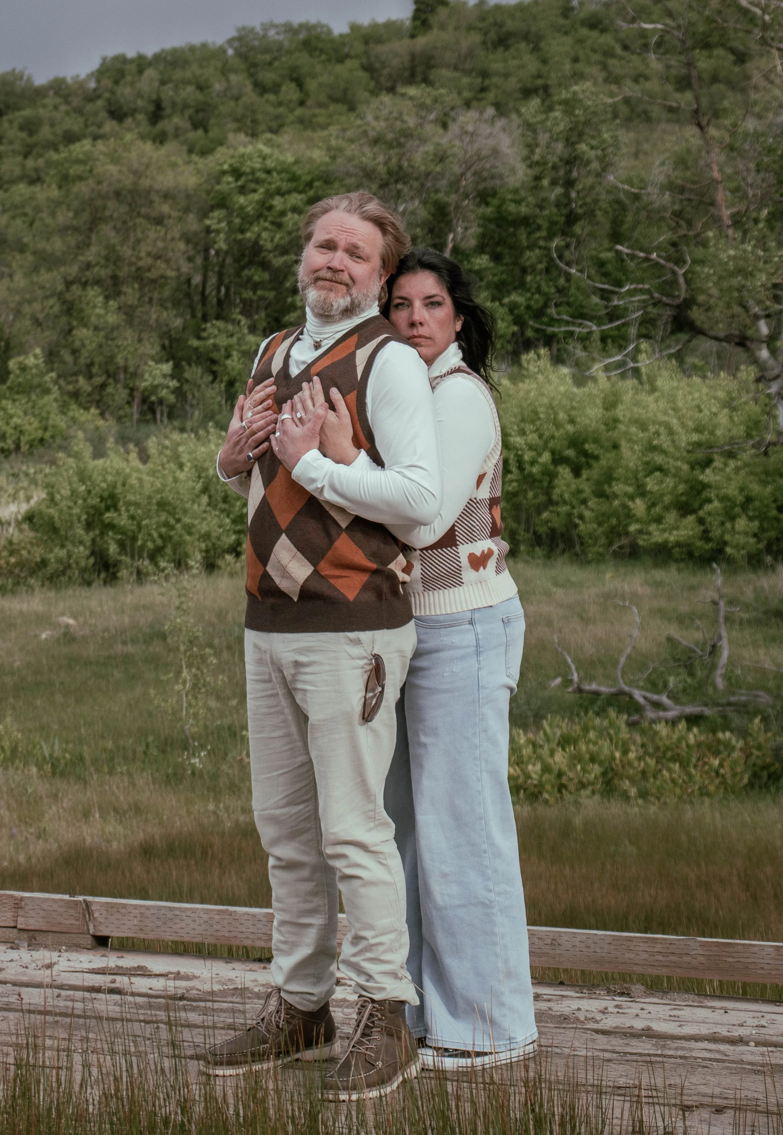 A man and a woman standing close together outdoors on a wooden walkway, embracing each other with their hands on their chests, with a background of green trees and a slightly cloudy sky.