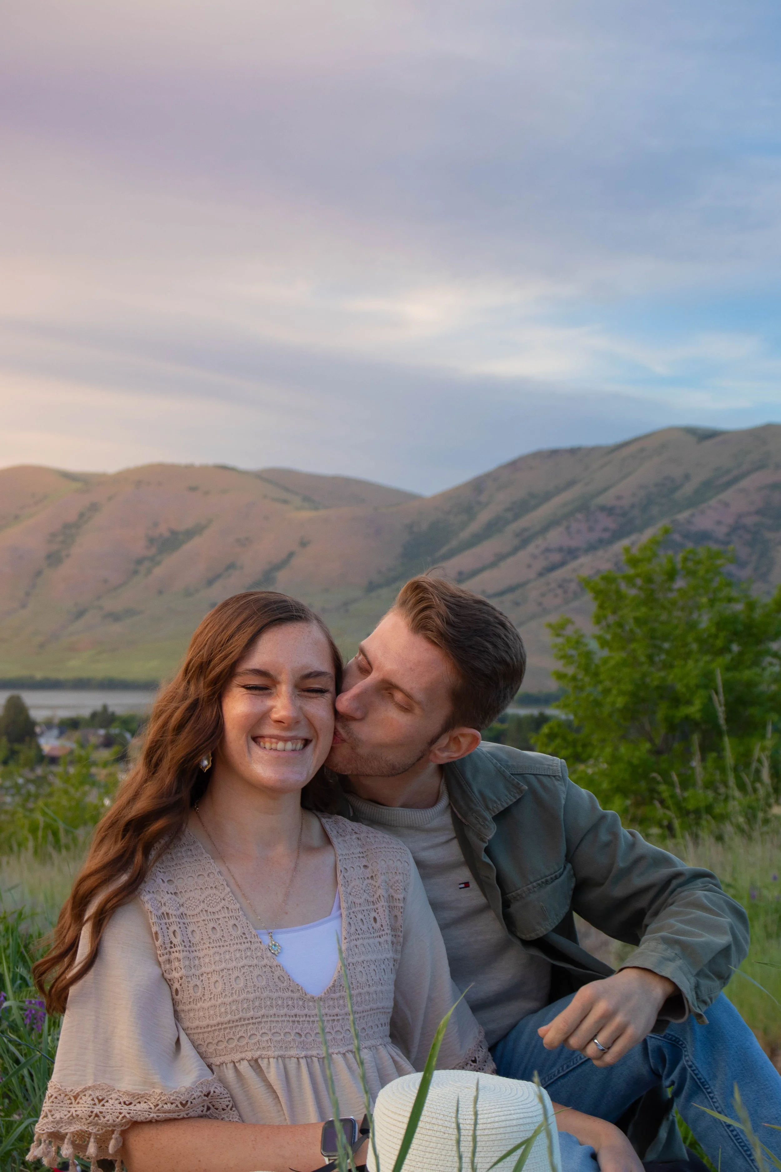 A young woman with long red hair is smiling with her eyes closed, while a young man next to her kisses her on the cheek in an outdoor setting with mountains and a cloudy sky in the background.