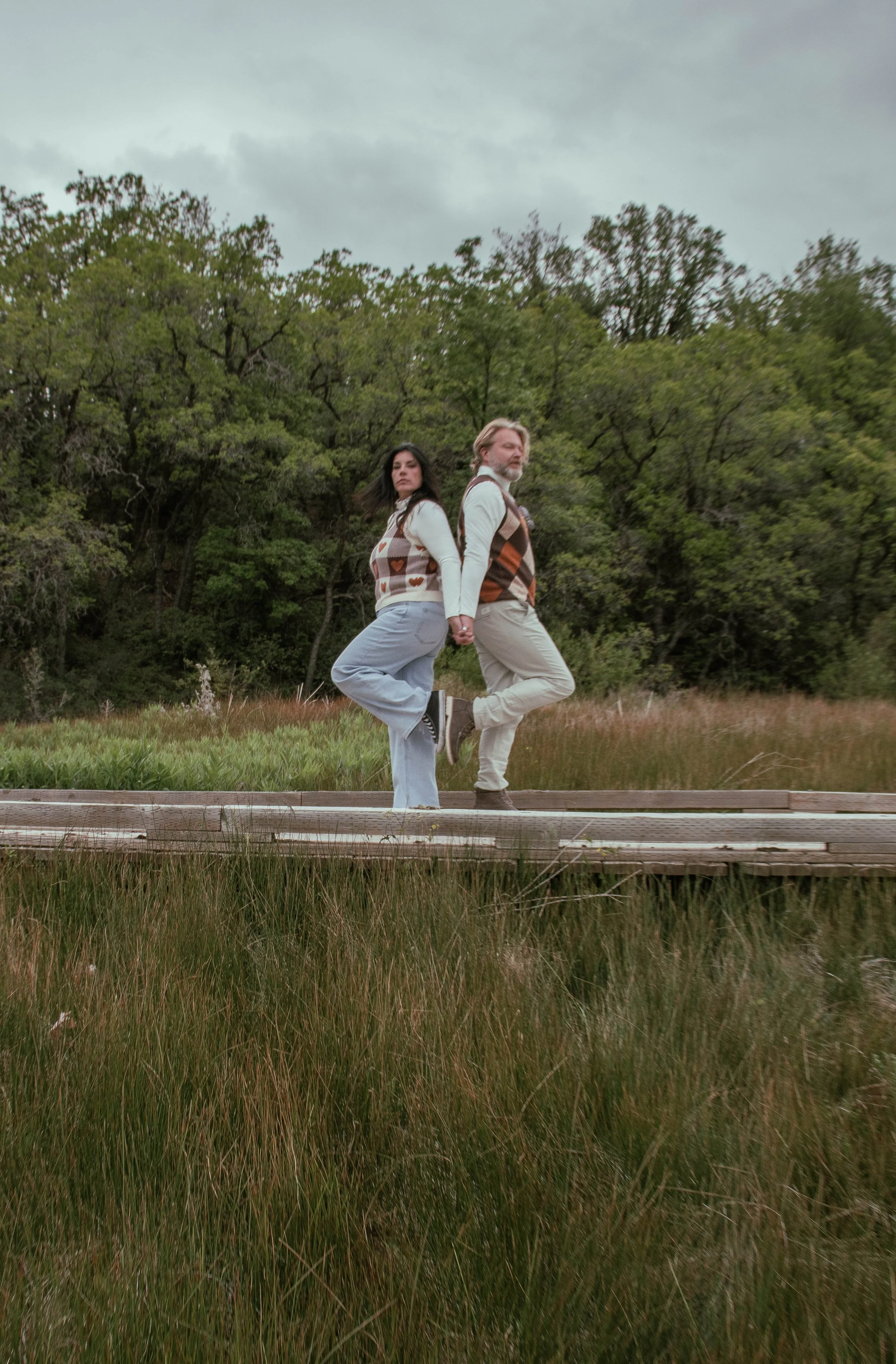 A couple holding hands, standing on a wooden dock, with a grassy field and trees in the background, on a cloudy day.