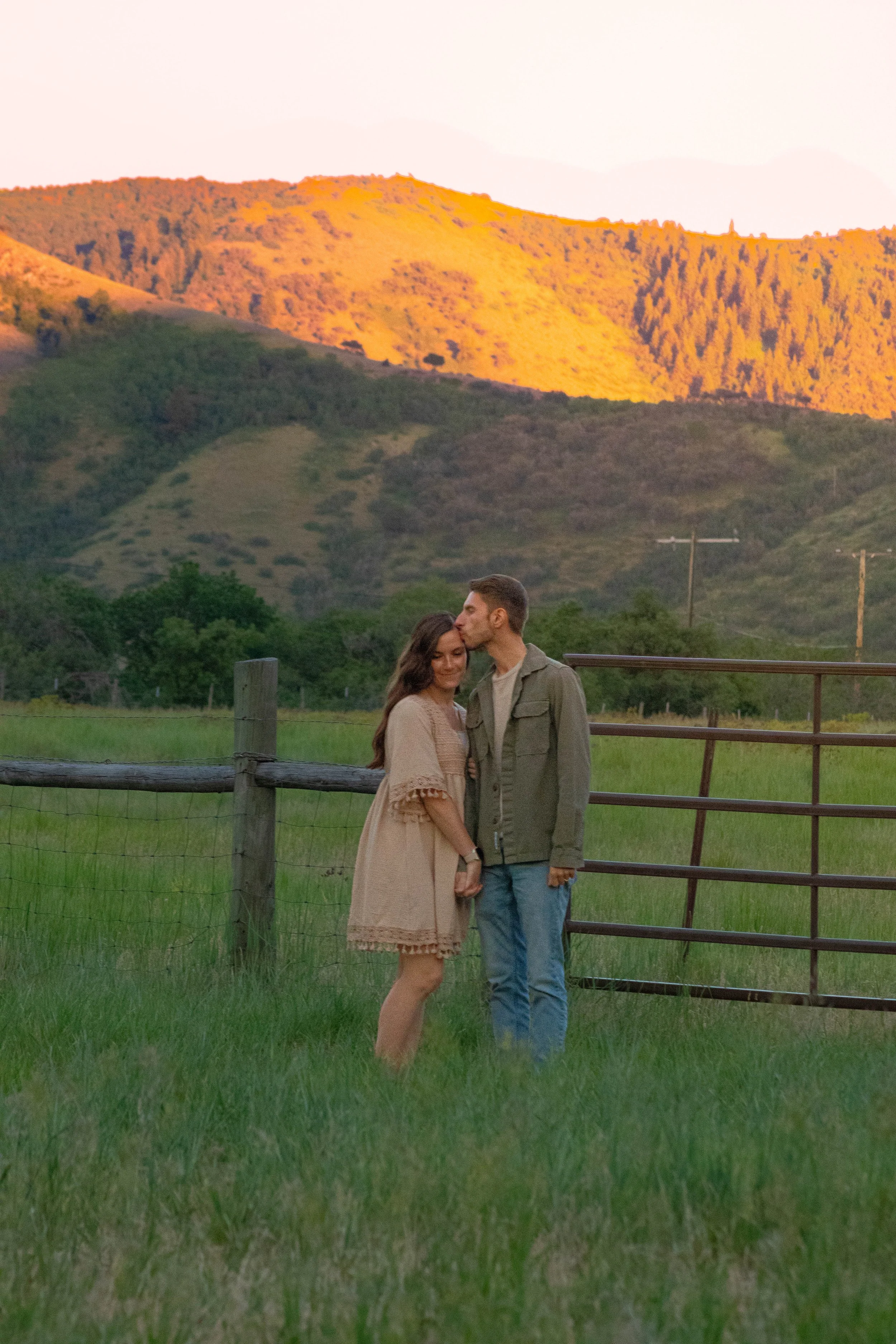 A couple stands together outdoors near a fence, with green grass and rolling hills in the background, illuminated by warm sunset light, mountains in the distance.