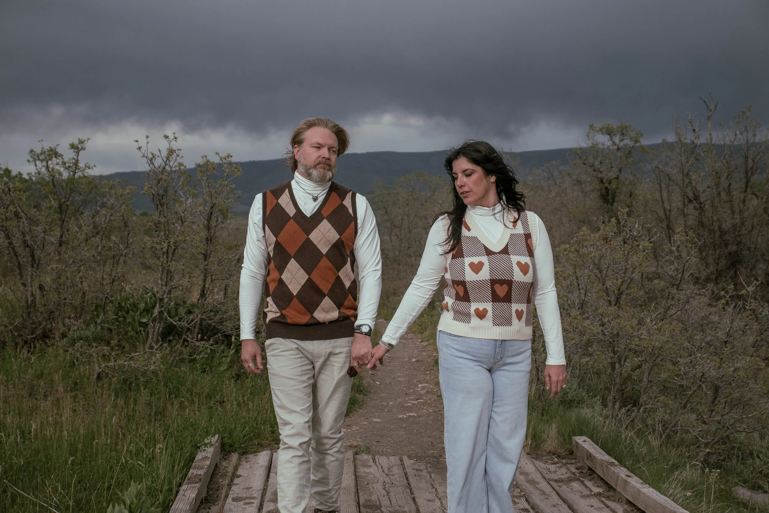 A man and a woman walking hand in hand on a wooden bridge outdoors under stormy clouds, surrounded by trees and hills.