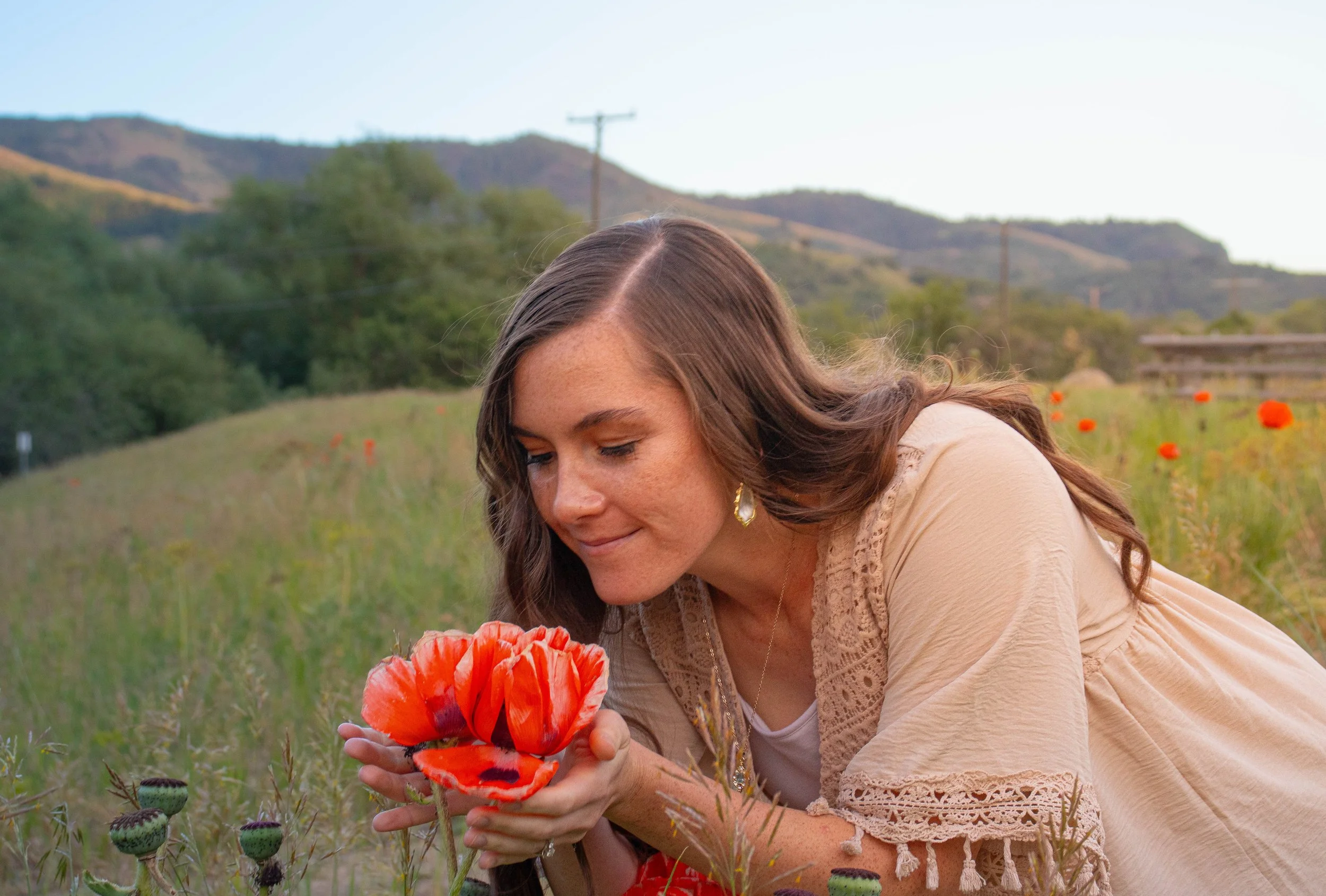 A woman with long brown hair, wearing a beige top with lace details, is holding a vibrant red poppy flower and looking at it in a field of poppies with mountains, trees, and a telephone pole in the background.