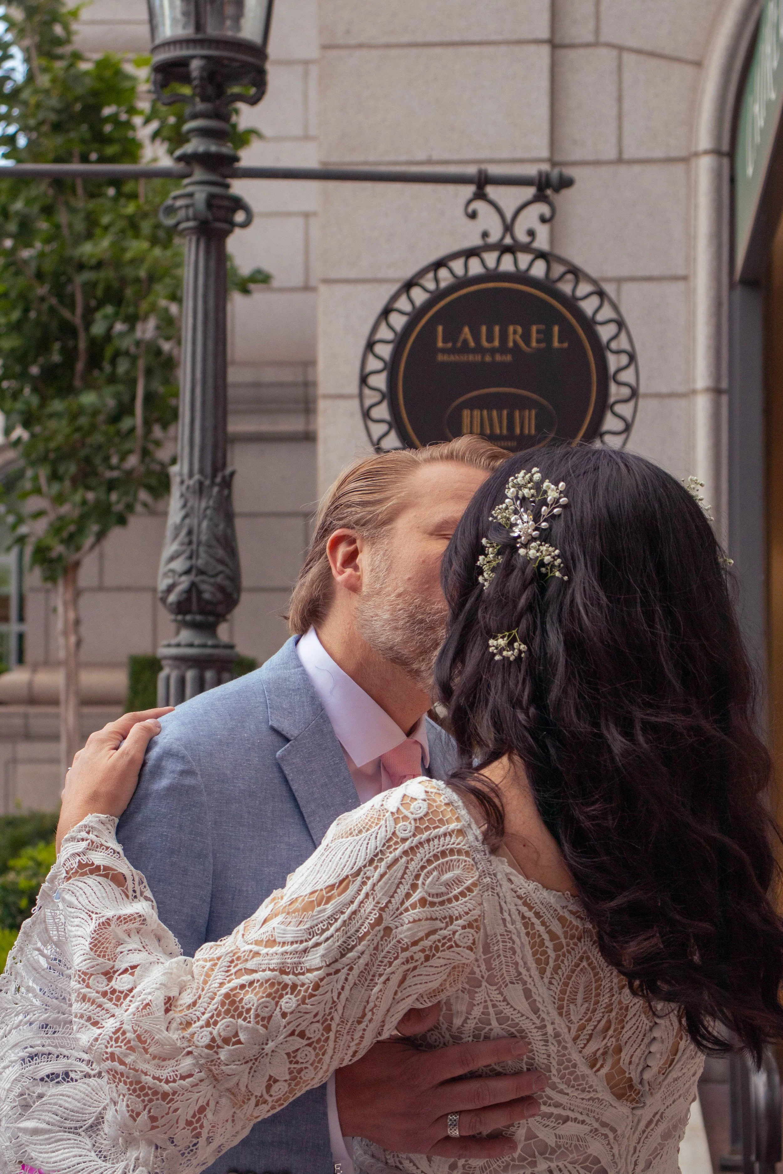A couple, dressed in wedding attire, closes their eyes as they kiss outdoors in front of a building with a sign that says 'Laurel Dining & Bar'.