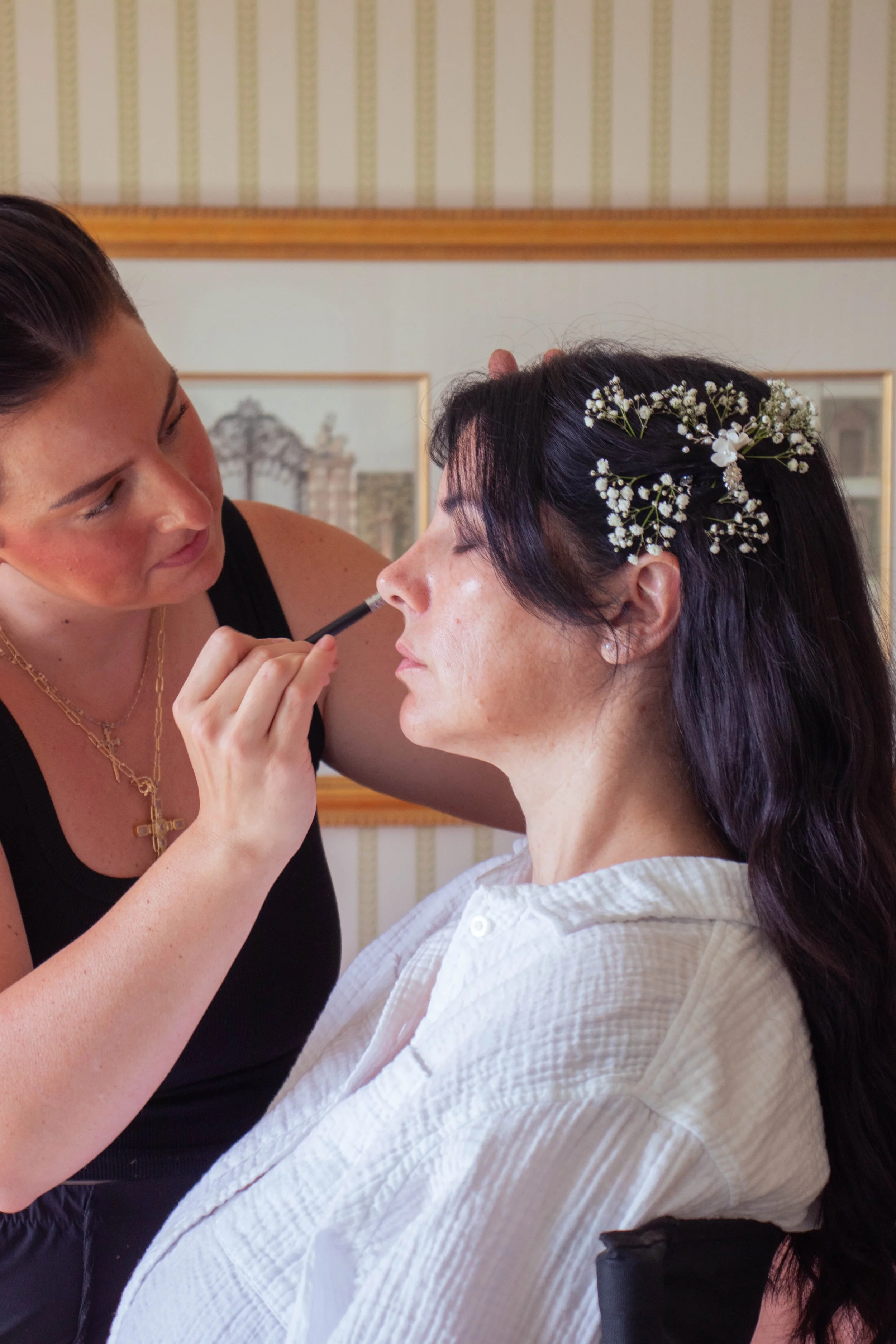 A woman with dark hair and floral hair accessory is getting her makeup done by a makeup artist wearing a black top and gold jewelry.