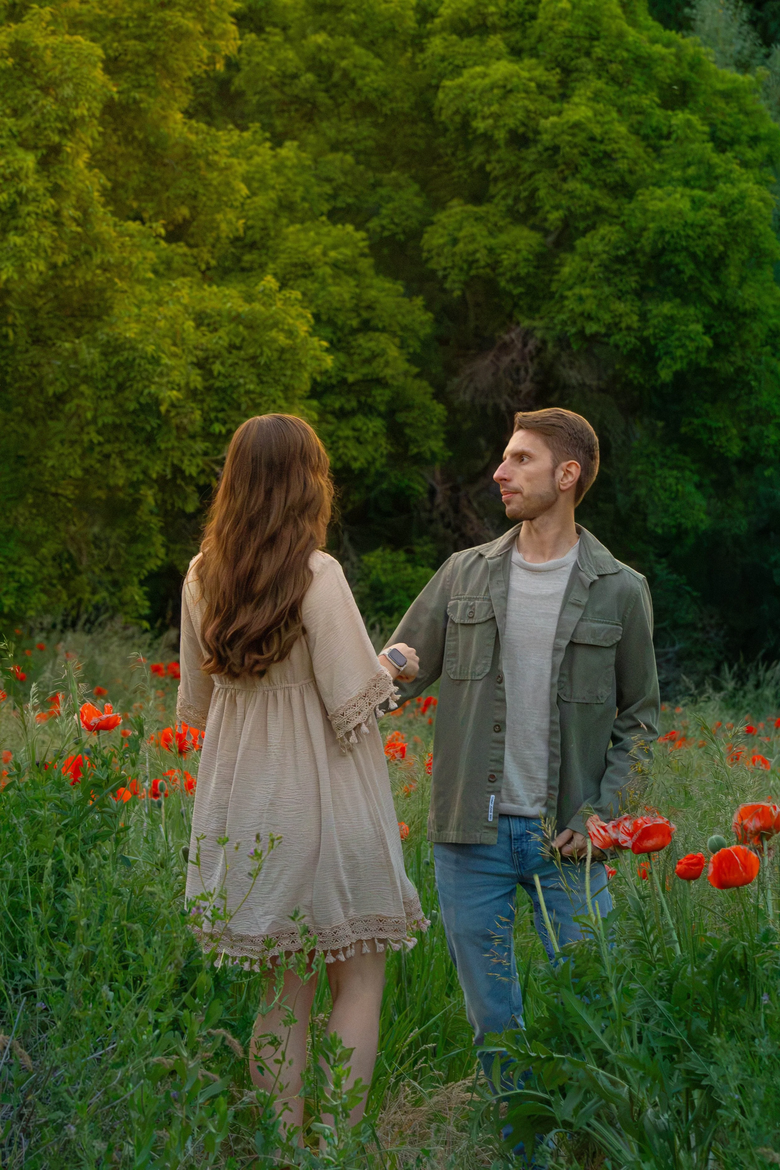A man and a woman standing in a field of red poppies, holding hands, with a backdrop of lush green trees.