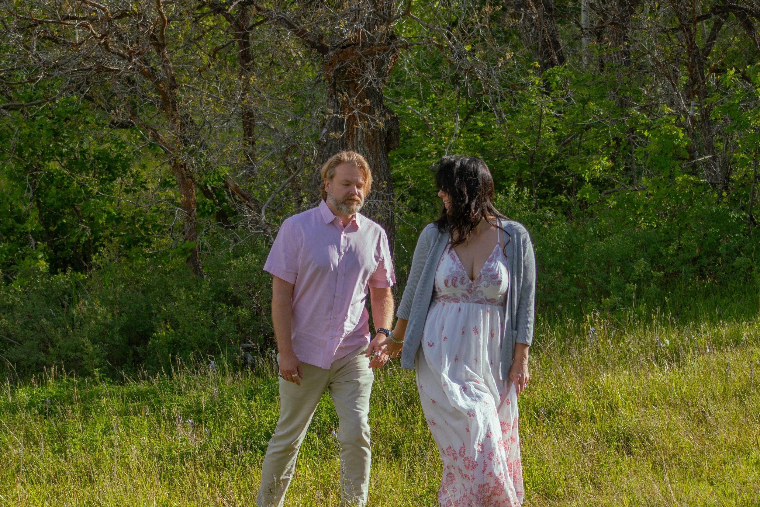 A man and woman holding hands walking through a grassy field with trees in the background.
