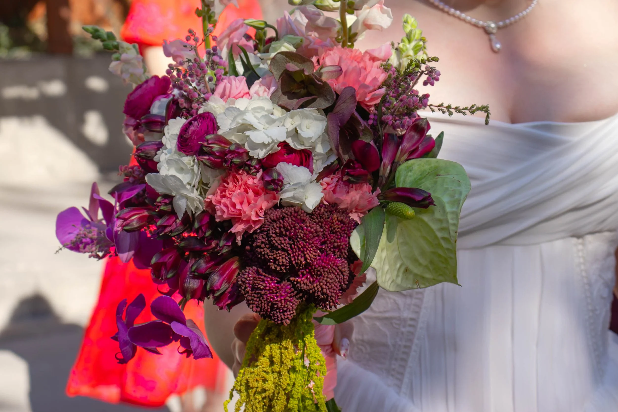 A woman in a white dress holding a colorful bouquet of pink, white, purple, and yellow flowers, with a pearl necklace around her neck.
