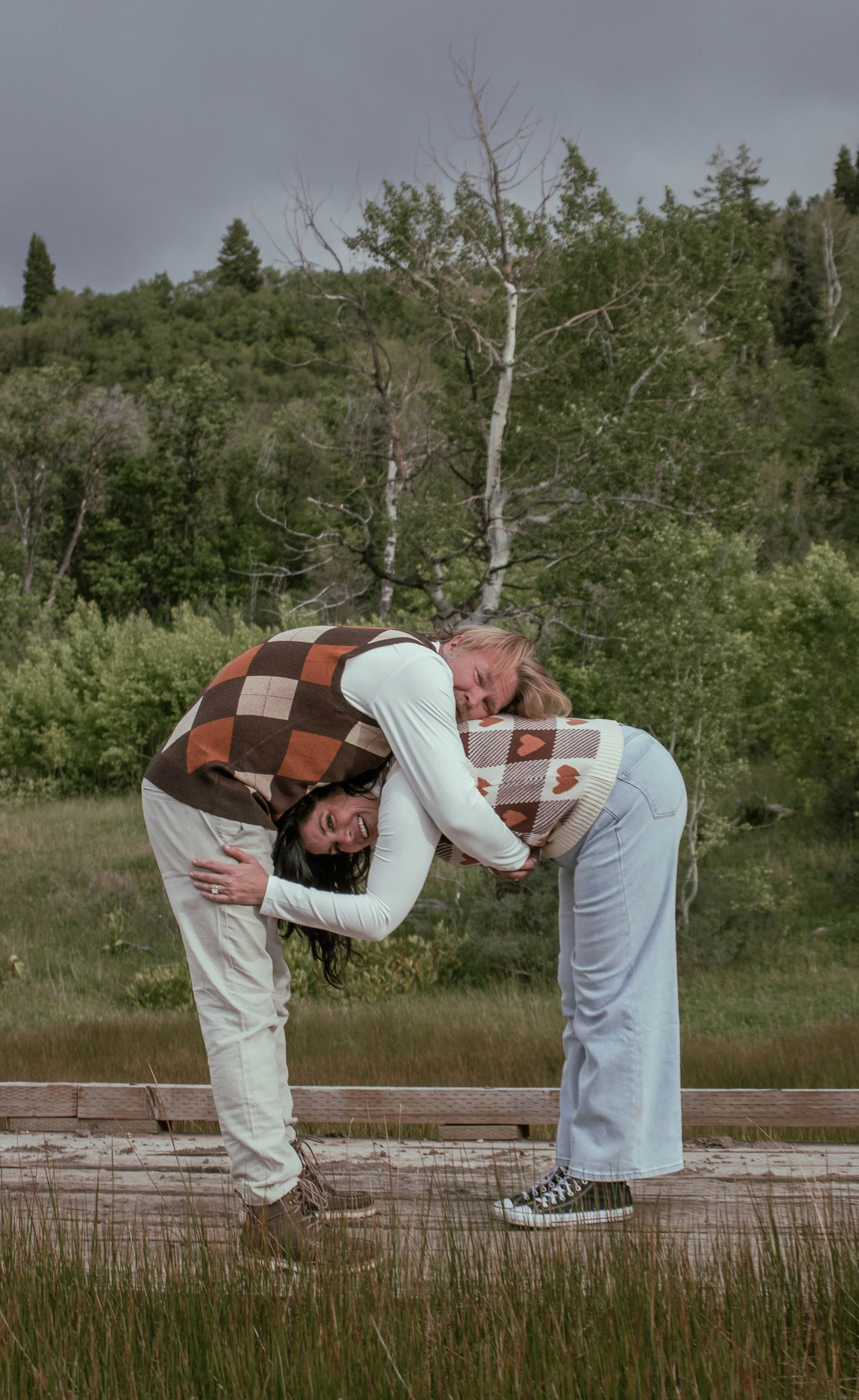 Two people, a man and a woman, are playing a game of human pretzel outdoors on a wooden path surrounded by grass and trees, with dark stormy clouds overhead.