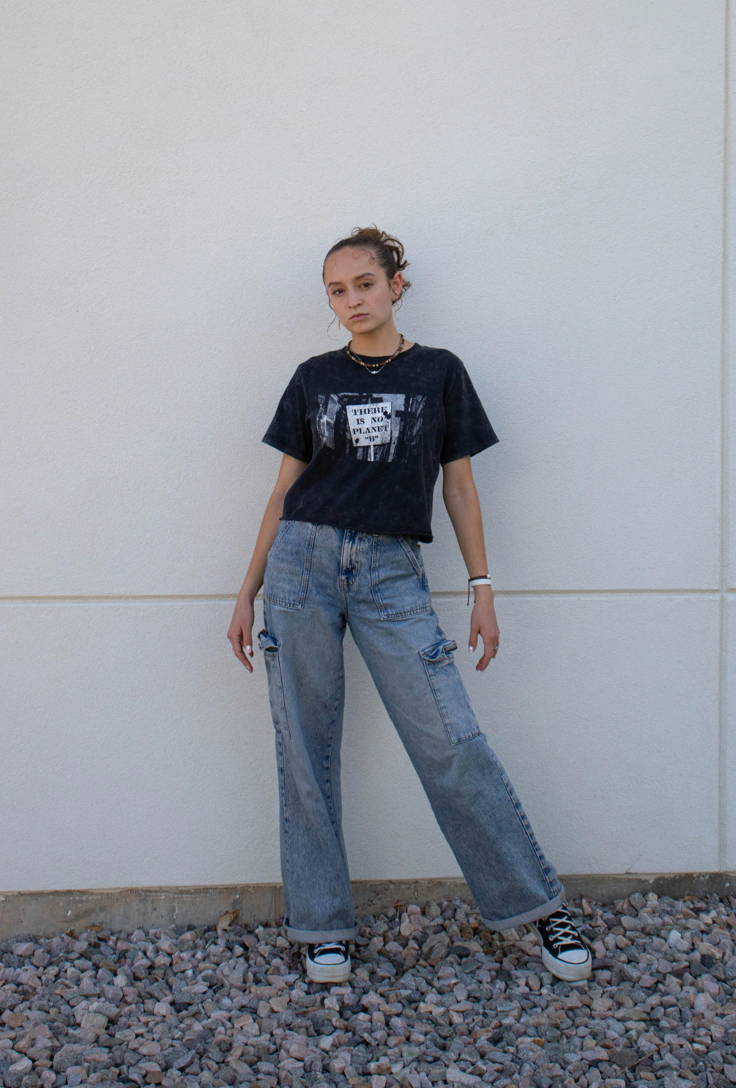 Young woman standing in front of a plain white wall, wearing a black graphic T-shirt, blue jeans, and sneakers, with her hair tied back and accessories around her neck.