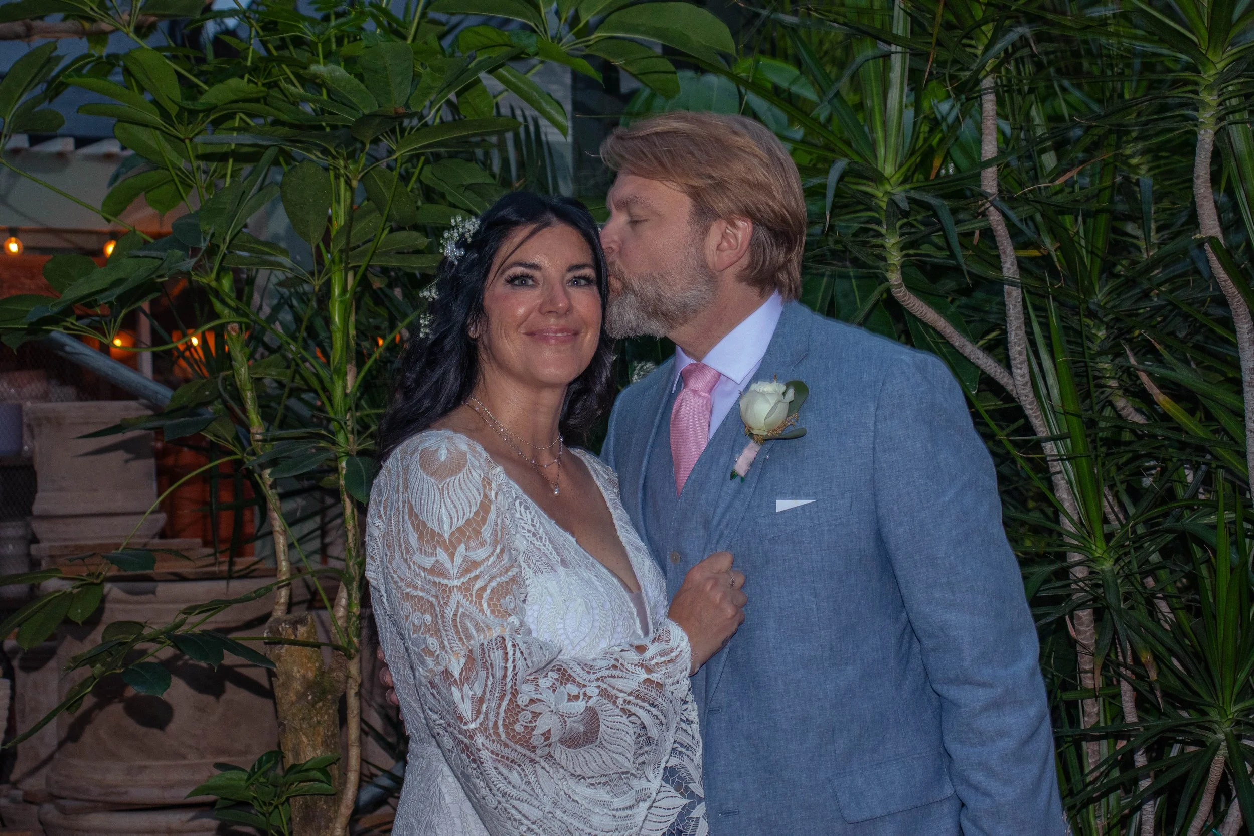 A couple, dressed in wedding attire, stands close together in front of green plants. The woman has dark hair and is wearing a white lace wedding dress, and the man has light hair and a beard, wearing a light blue suit with a pink tie and a boutonnièr
