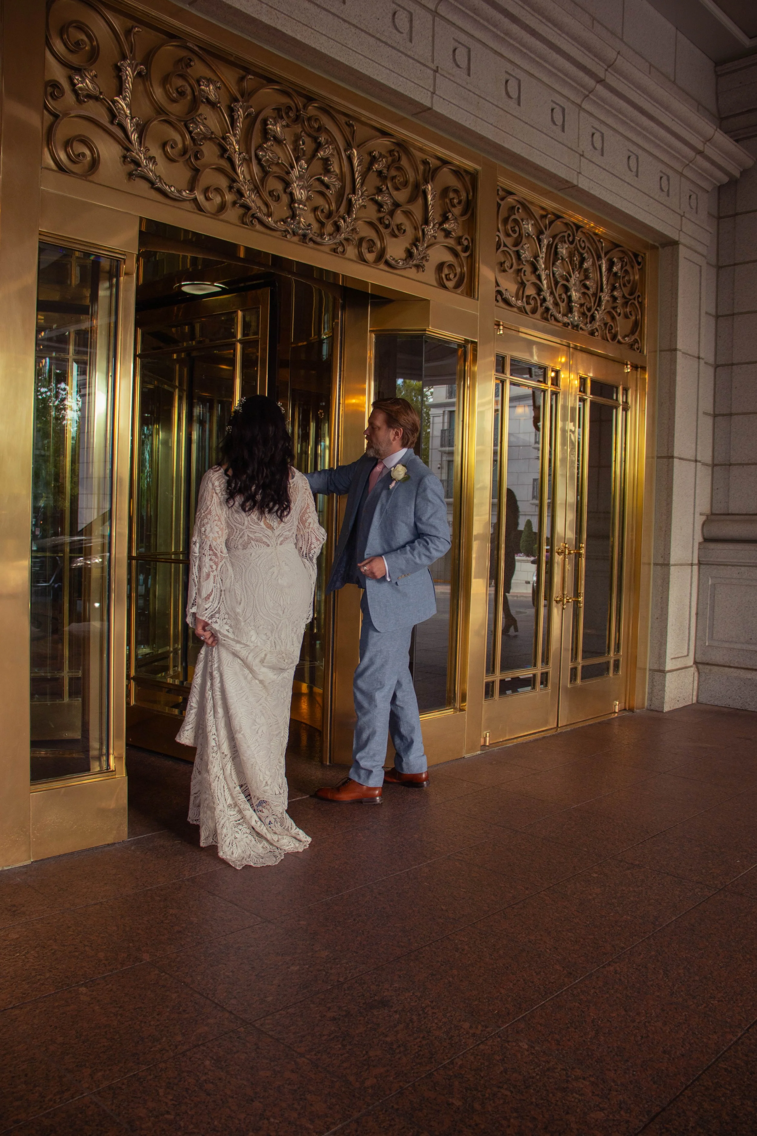 A bride and groom standing at the entrance of a building with ornate gold doors and intricate gold decorative elements above the doors, sharing a moment together.