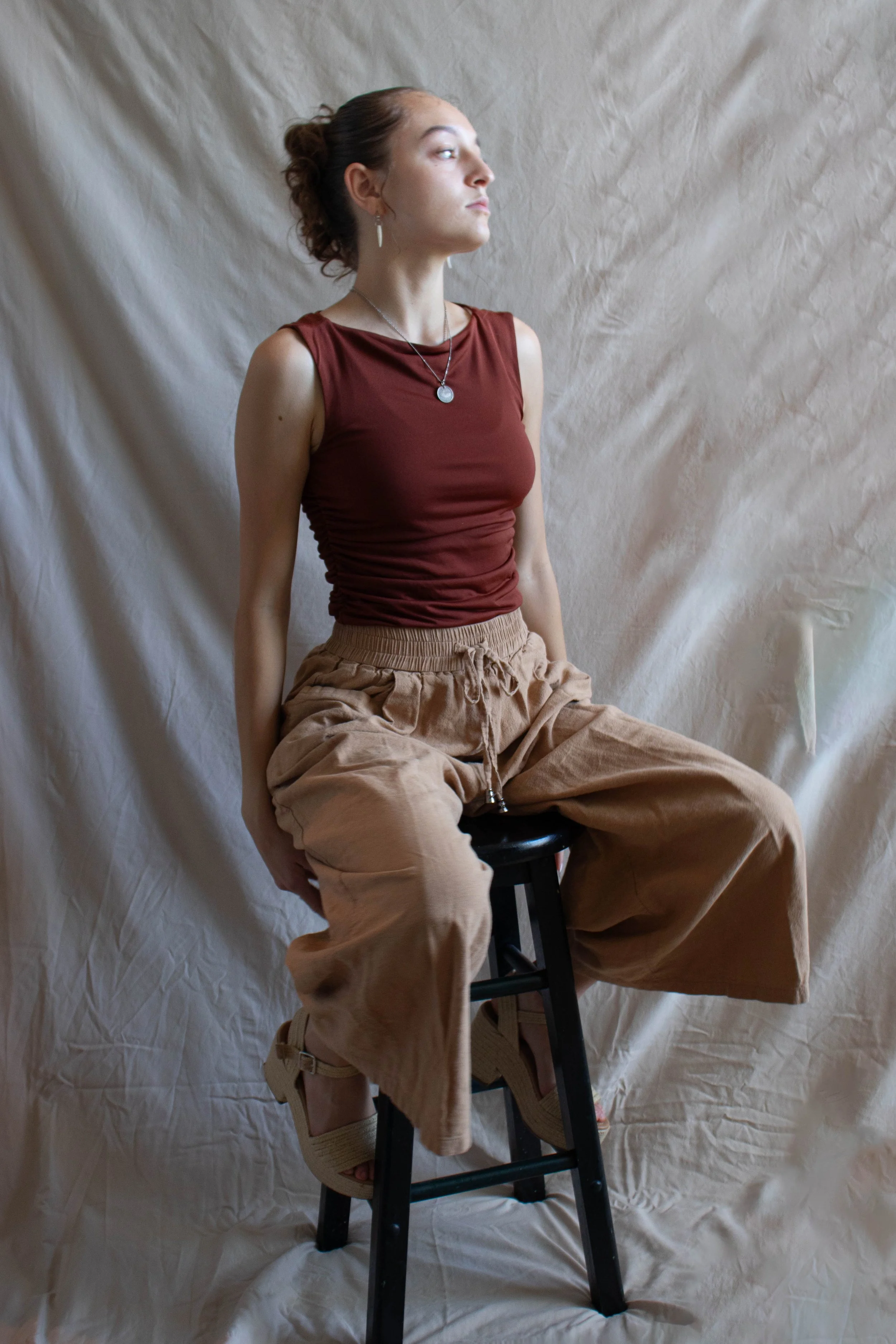 A young woman with brown hair tied back, wearing a sleeveless maroon top and tan cargo pants, sits on a black stool against a crumpled beige fabric backdrop. She is looking to her left with a calm expression.