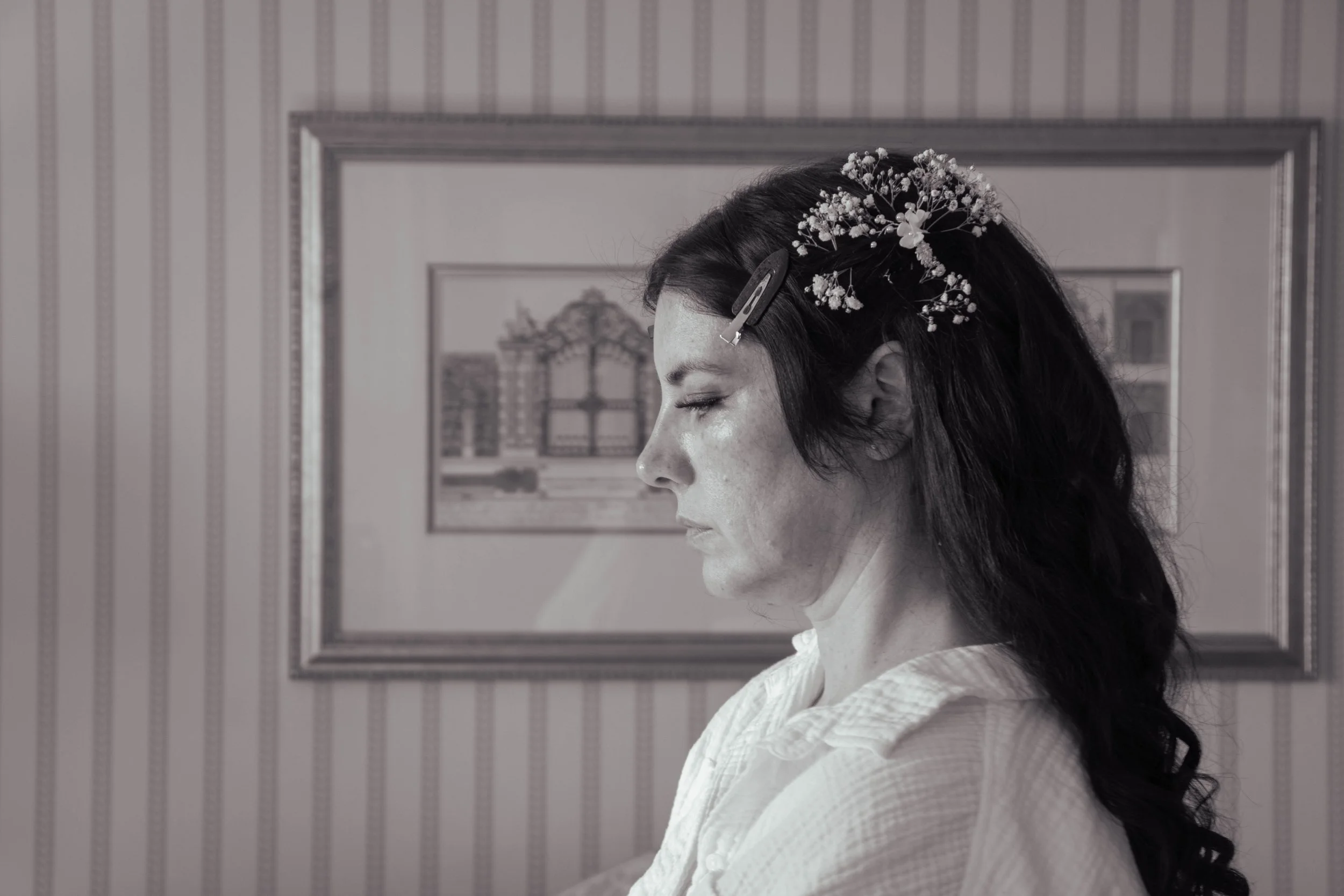 Black and white photo of a woman with dark hair, styled with small white flowers and hair clips, wearing a white textured blouse, standing in front of a framed picture and striped wallpaper.