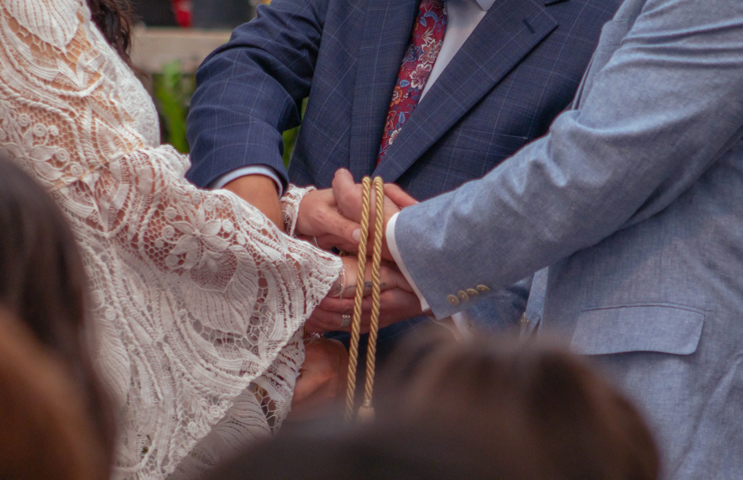 A close-up of two people holding hands during a wedding ceremony, with the focus on the hands and the wedding rings, and the bride wearing a lace dress and the groom in a suit with a floral tie.
