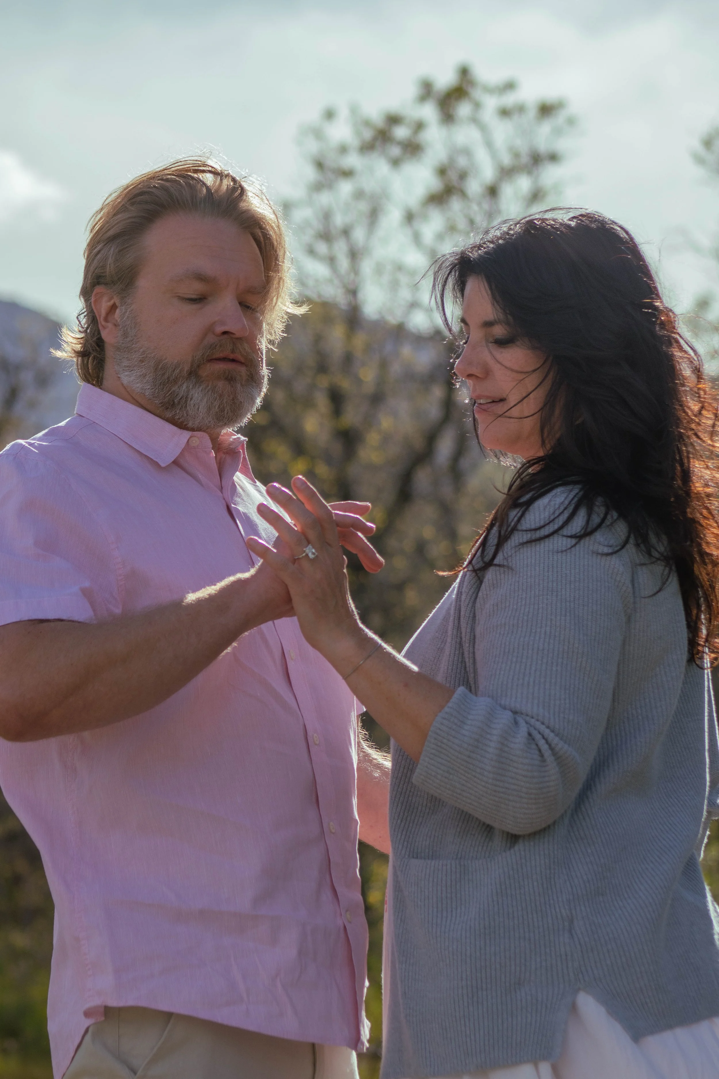 A man with a beard and a woman with long dark hair stand close together outdoors, holding hands and gazing at each other, with trees and a partly cloudy sky in the background.