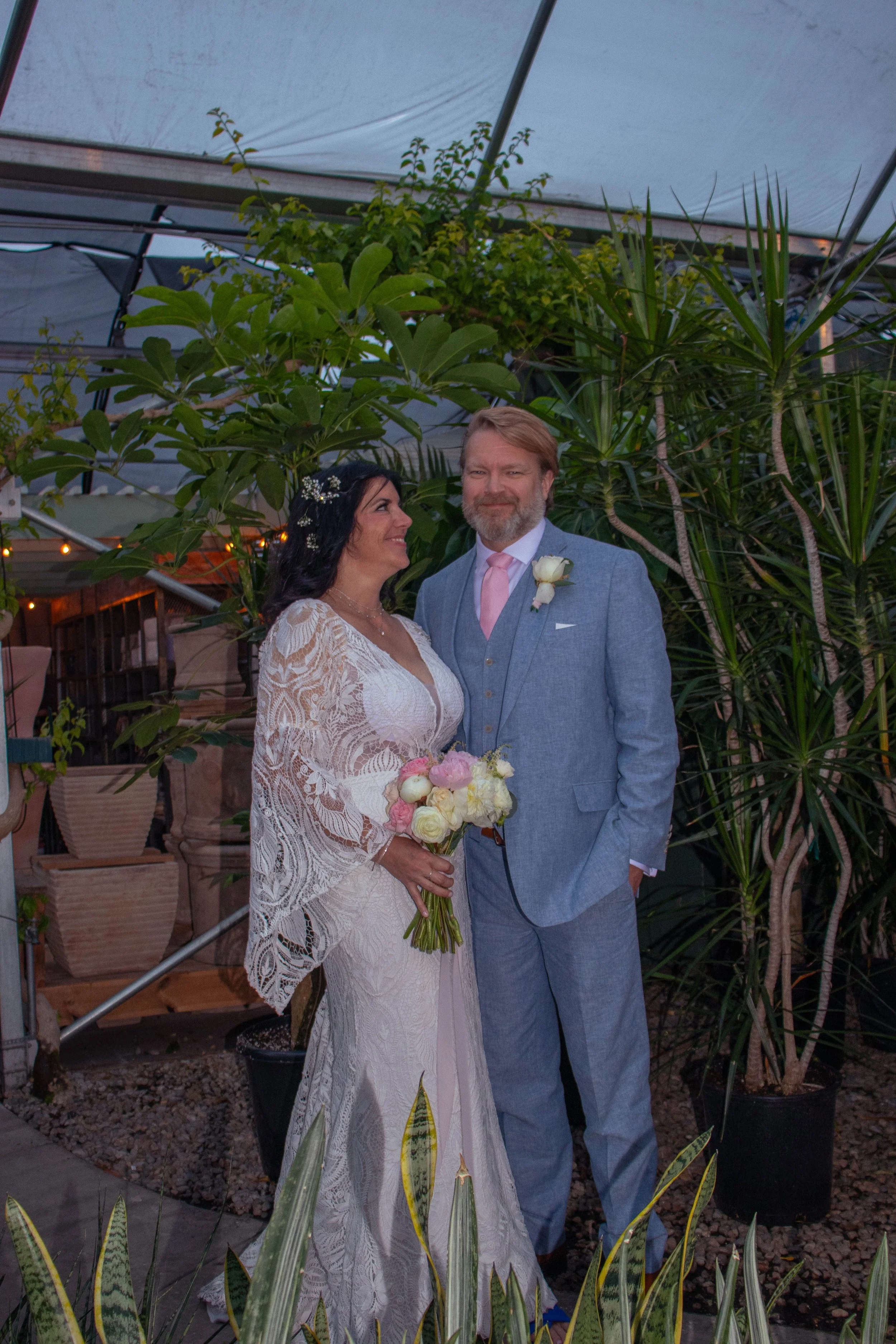A bride and groom standing together outdoors at a wedding, surrounded by plants and trees, with the bride holding a bouquet of flowers.