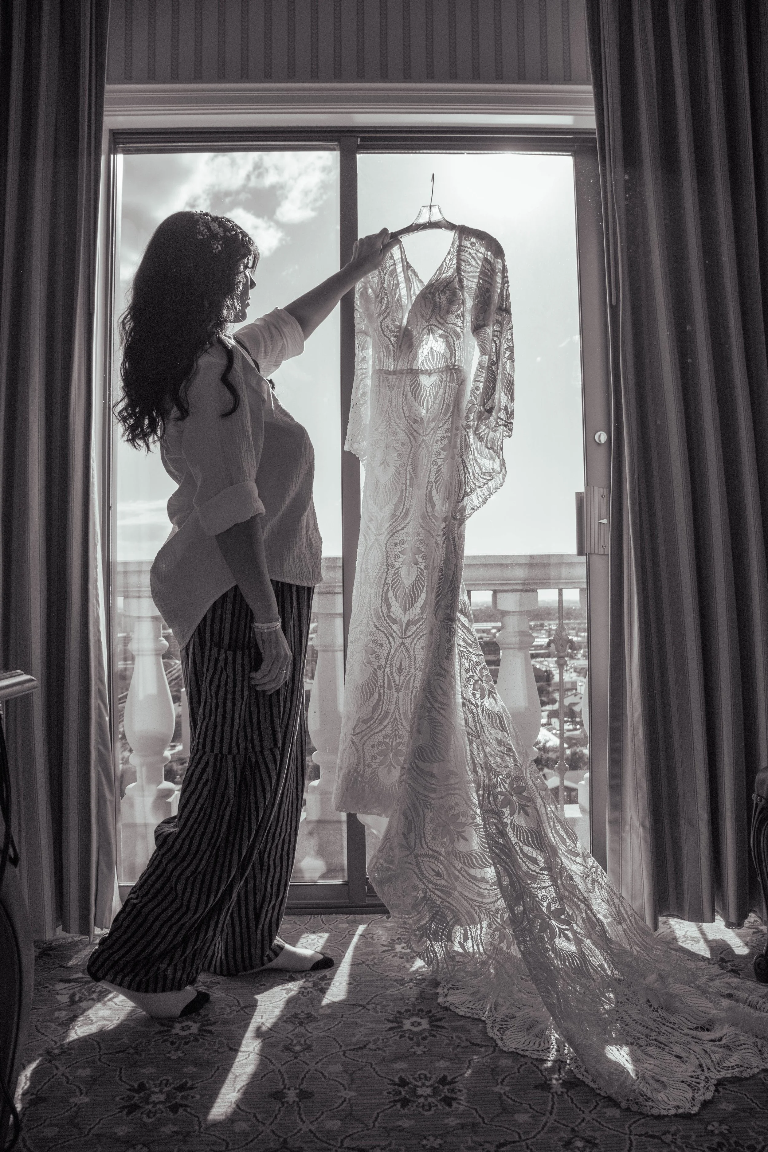 A woman with long hair, wearing striped pants and a loose shirt, stands inside looking at a lace wedding dress hanging on a hanger in front of her. The dress is backlit by sunlight, with a balcony and cloudy sky visible through the window behind it.