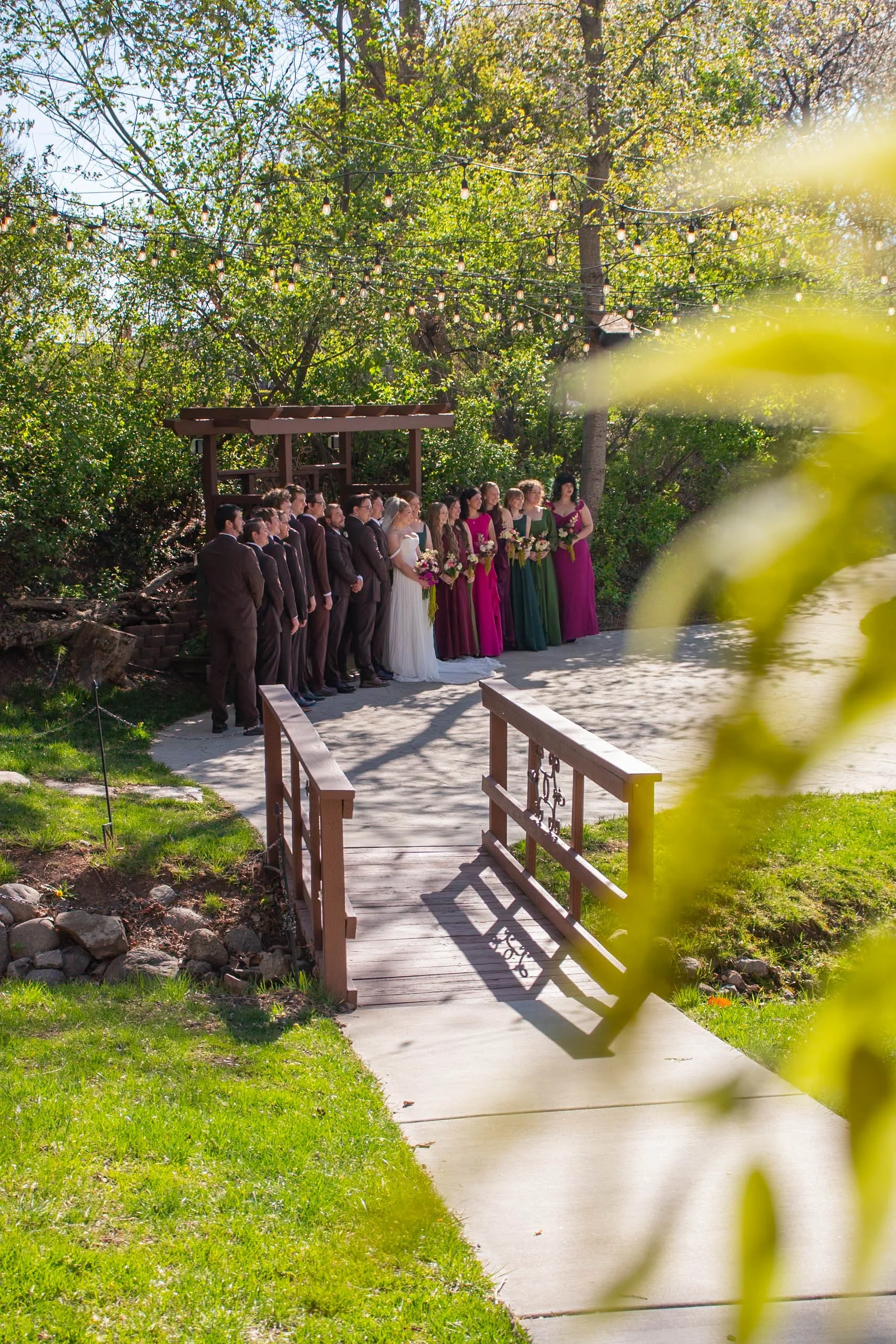 Group of people standing outdoors on a wedding ceremony, with trees and string lights hanging overhead, during daytime.