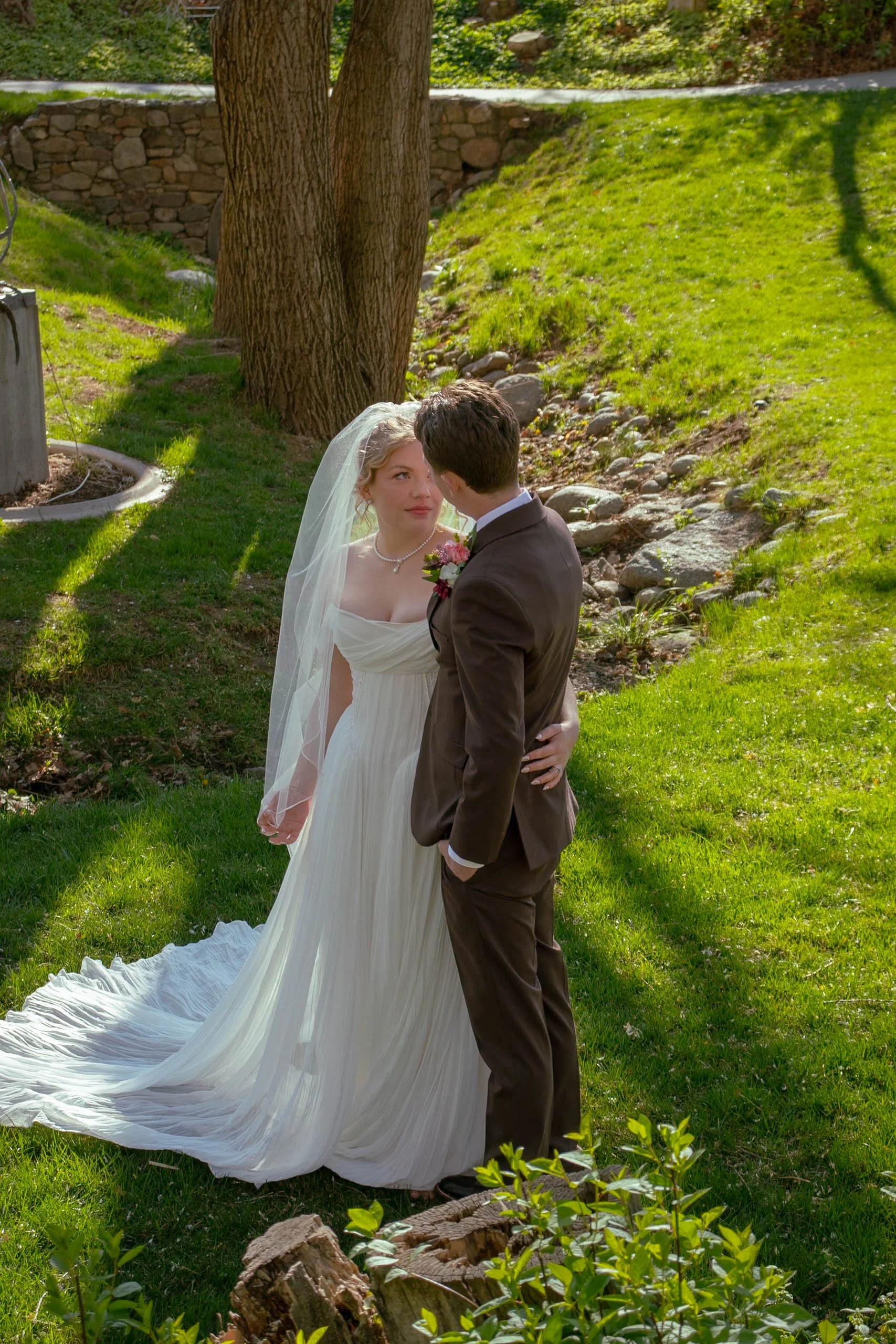 A bride and groom embracing outdoors on a sunny day in a garden with green grass, trees, and a stone wall, sharing an intimate moment.