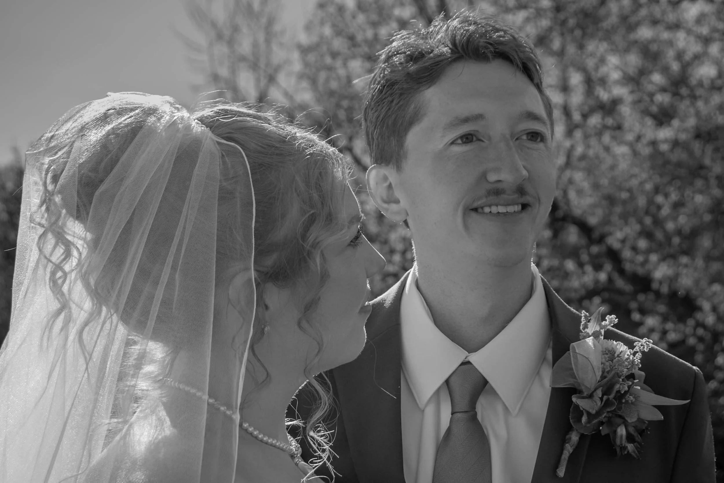A black-and-white photograph of a young couple, likely on their wedding day. The bride, with curly hair partially covered by a veil, is affectionately leaning toward the groom. The groom has a short haircut, light facial hair, and is dressed in a sui