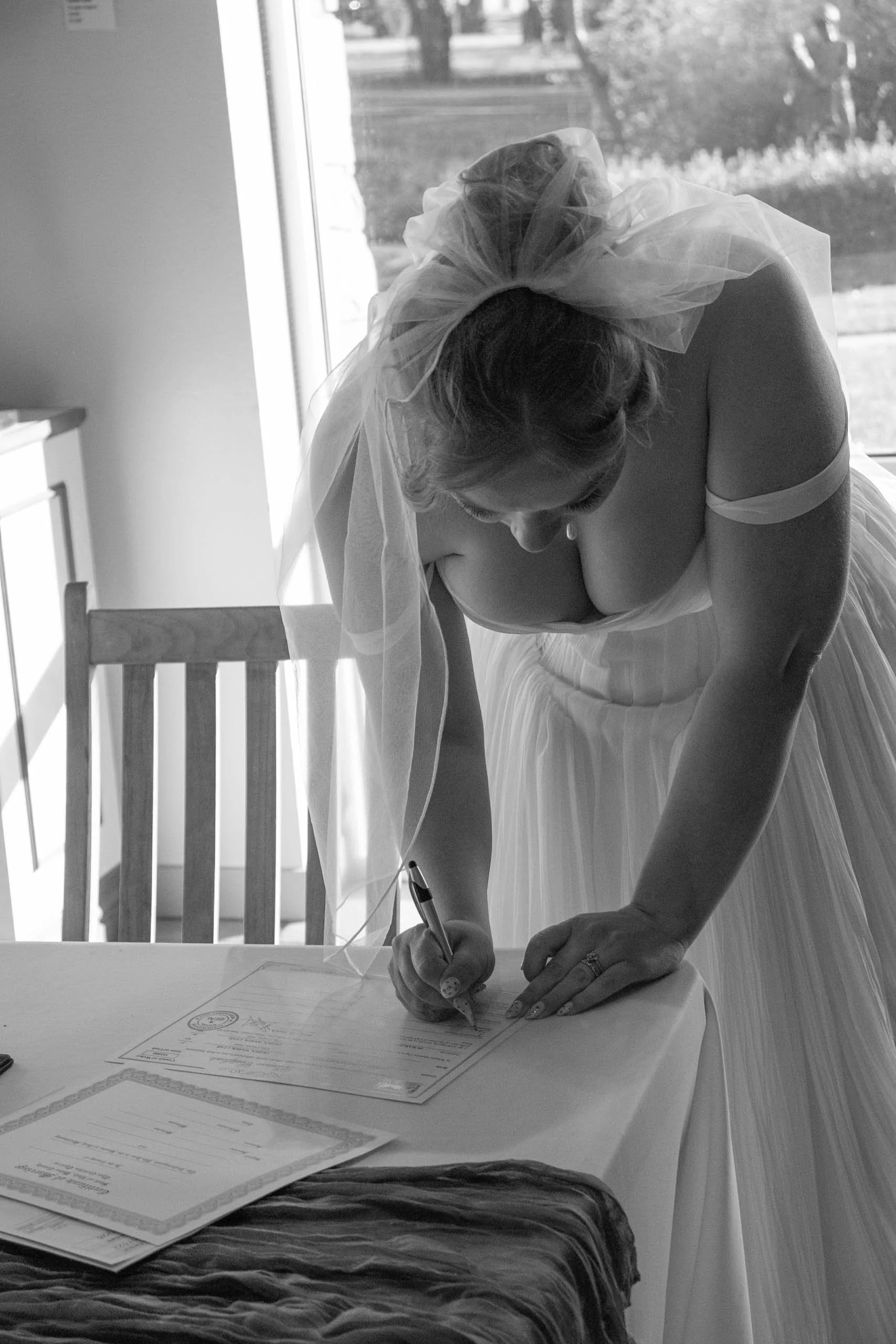 A woman wearing a wedding dress signs a document at a table with certificates. She is wearing a veil with a bow and is in a room with from a window showing trees outside.