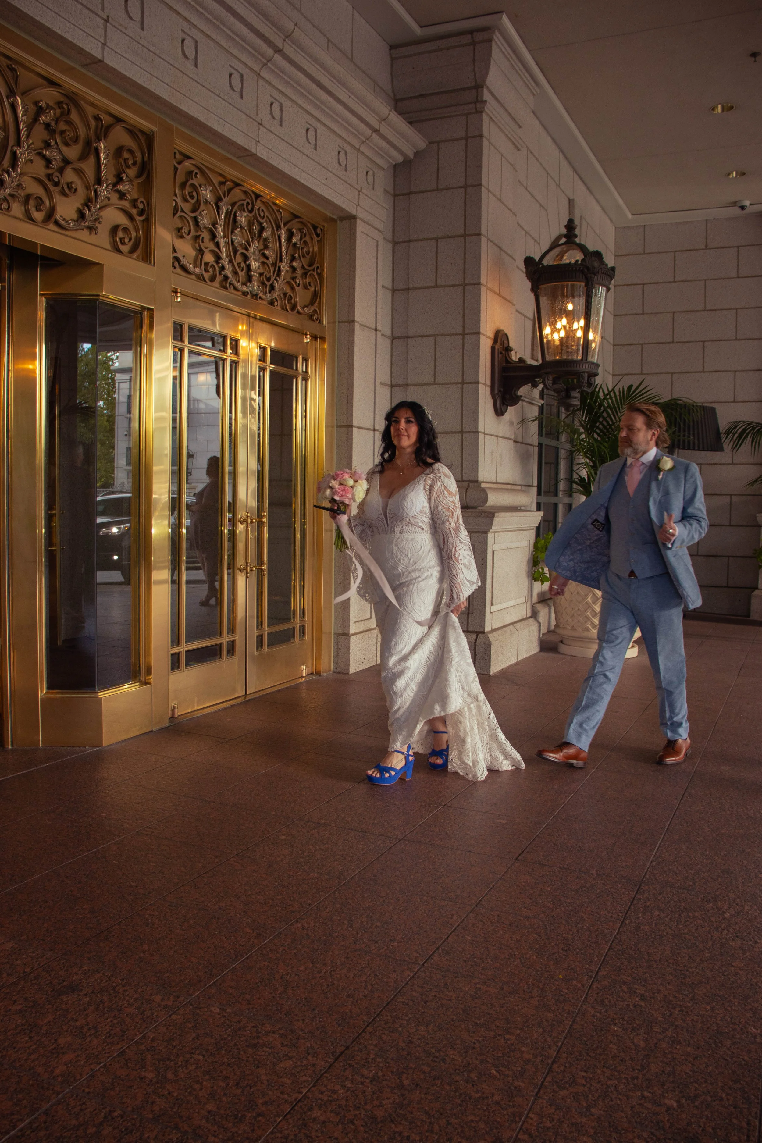 A woman in a white wedding dress holding a bouquet of flowers walking out of a building with a man in a light blue suit behind her.