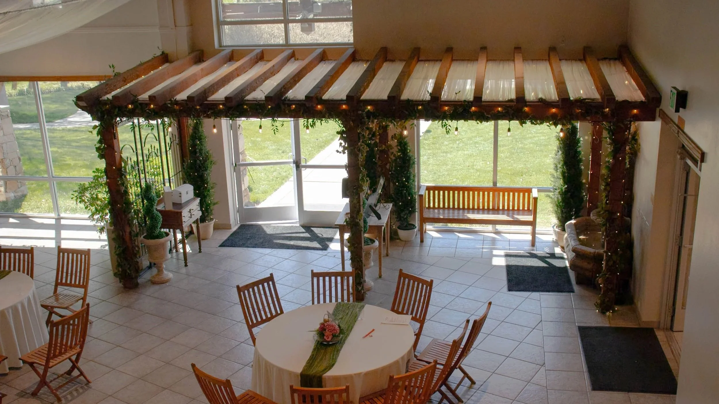Decorative entryway with a wooden archway adorned with greenery and string lights, glass doors, potted plants, and a wooden bench, leading outside to a grassy area.