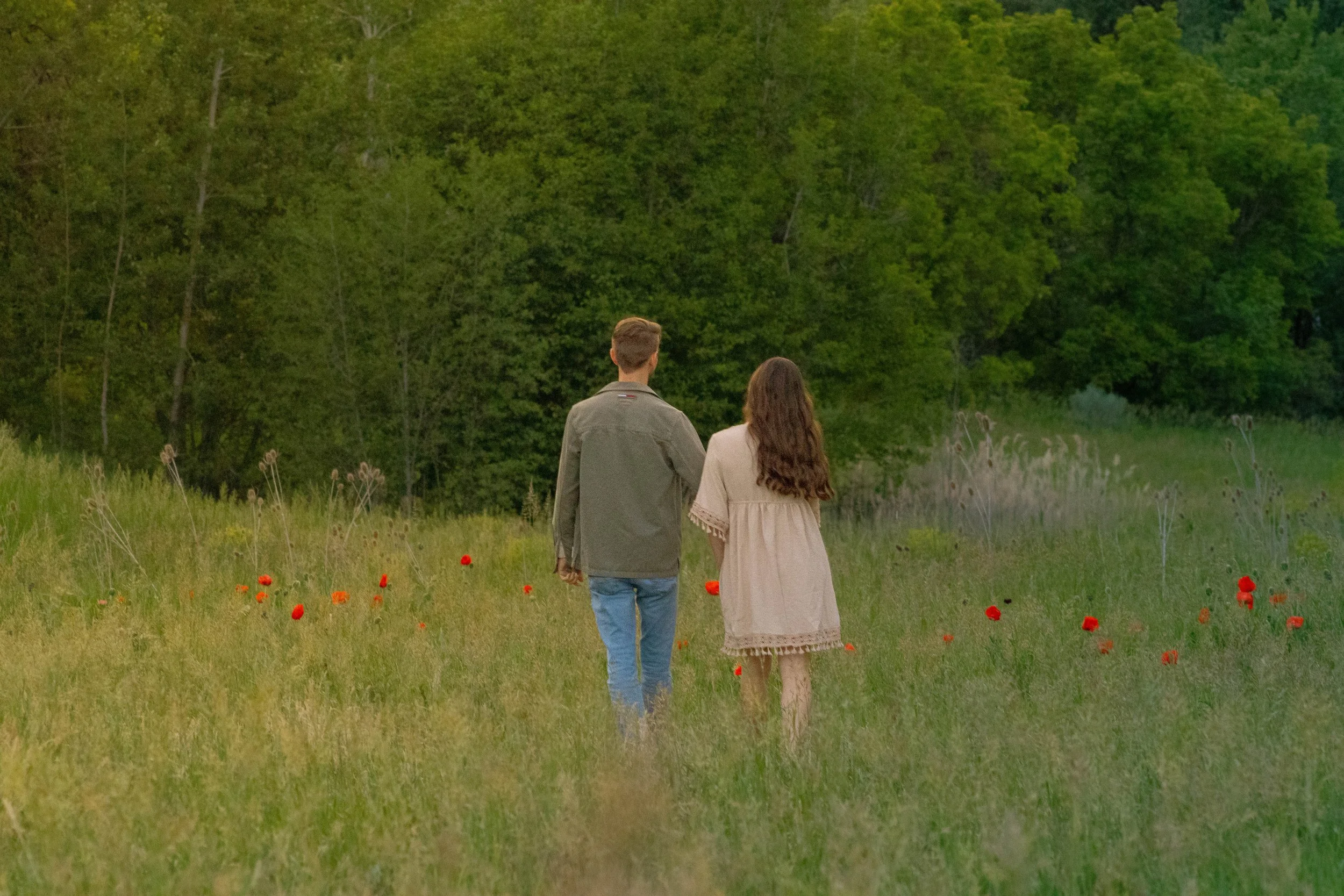 A couple walking hand in hand through a field with red poppies, surrounded by green trees.