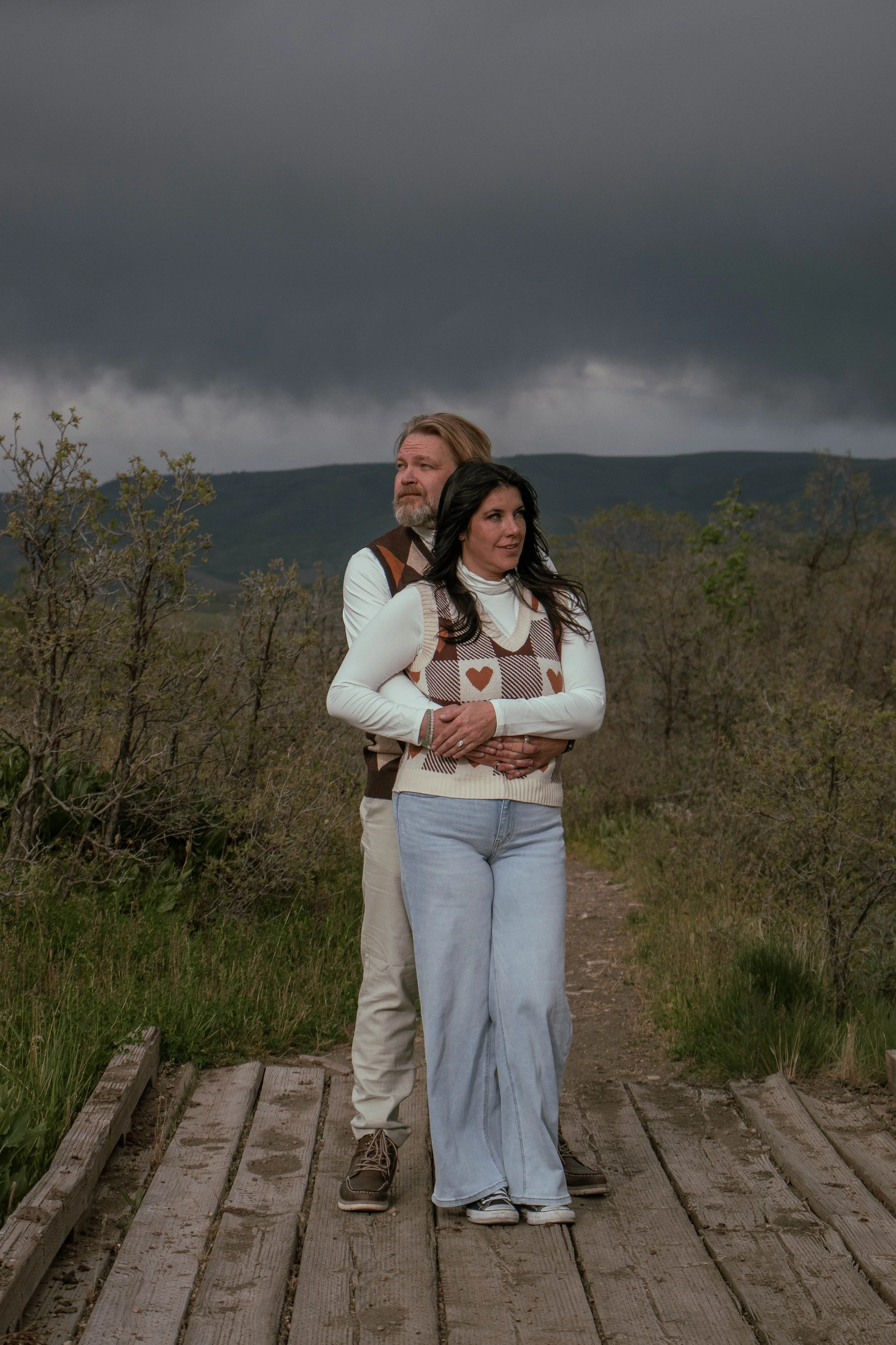 A man and a woman standing on a wooden bridge outdoors during a stormy day with dark, heavy clouds overhead and a landscape of trees and hills in the distance.