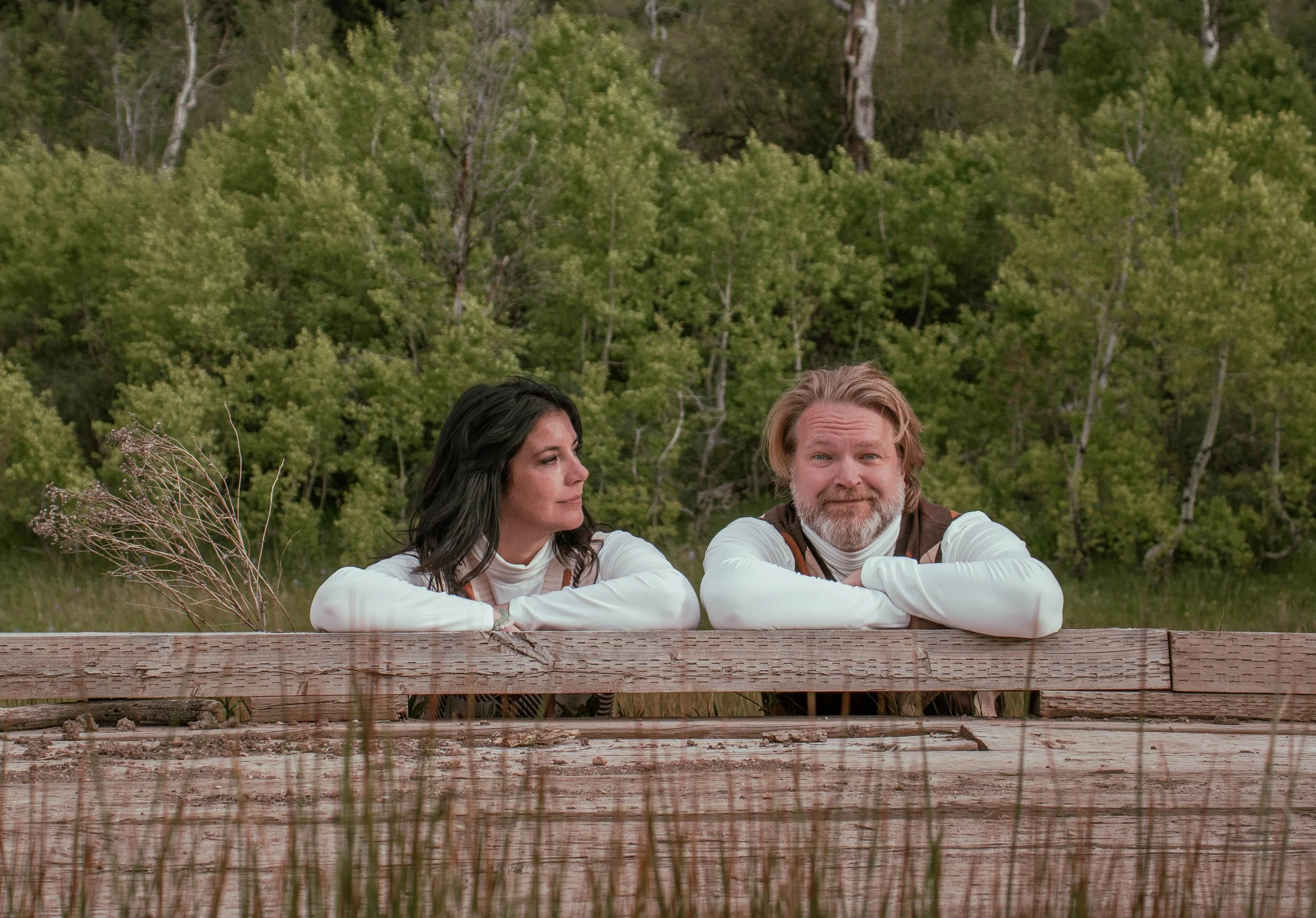 A man and a woman are sitting at a wooden picnic table outdoors, with green trees in the background. They are resting their arms on the table, with the woman looking at the man and the man looking directly at the camera.