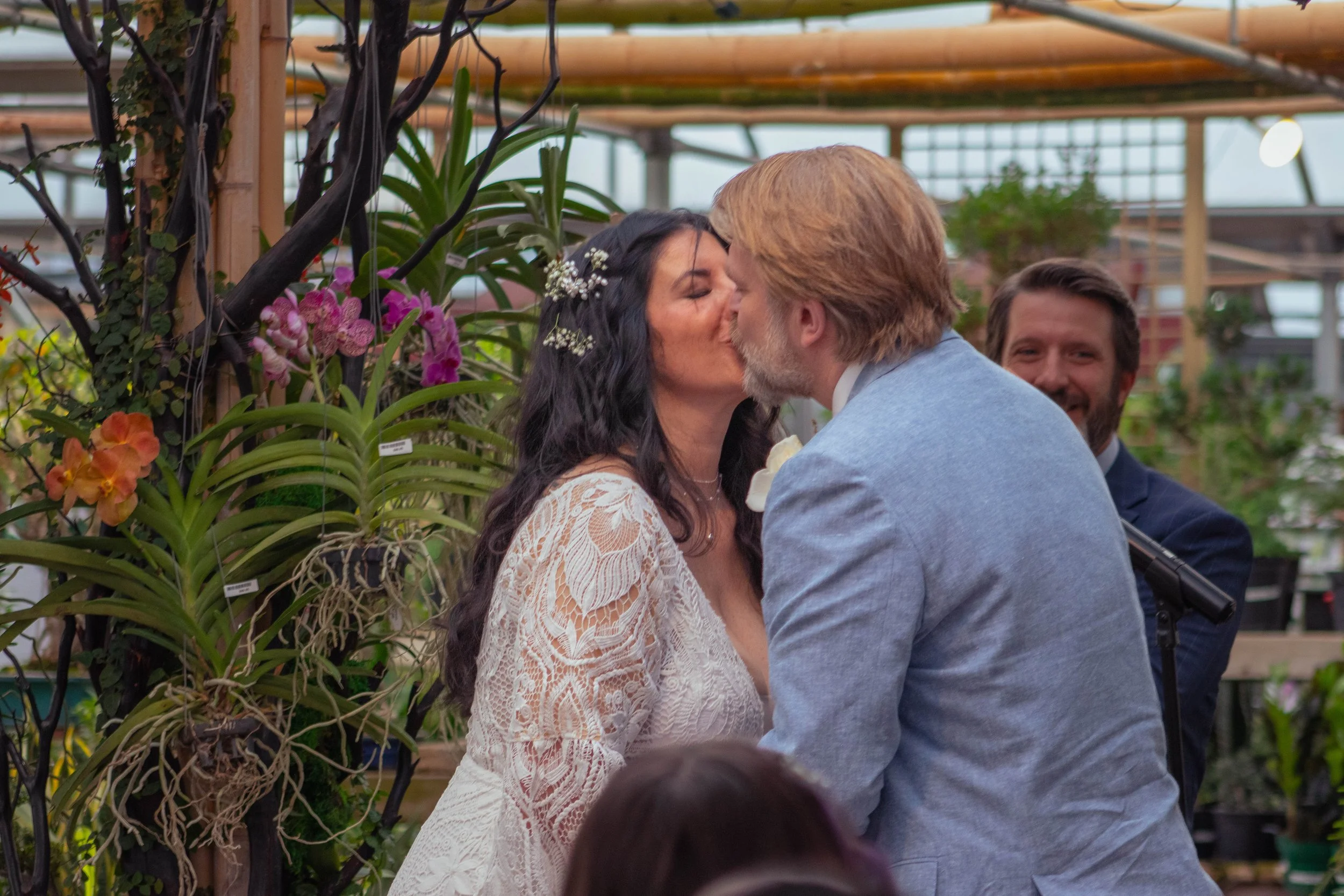 A couple kissing each other in a greenhouse surrounded by orchids, with a man smiling in the background.