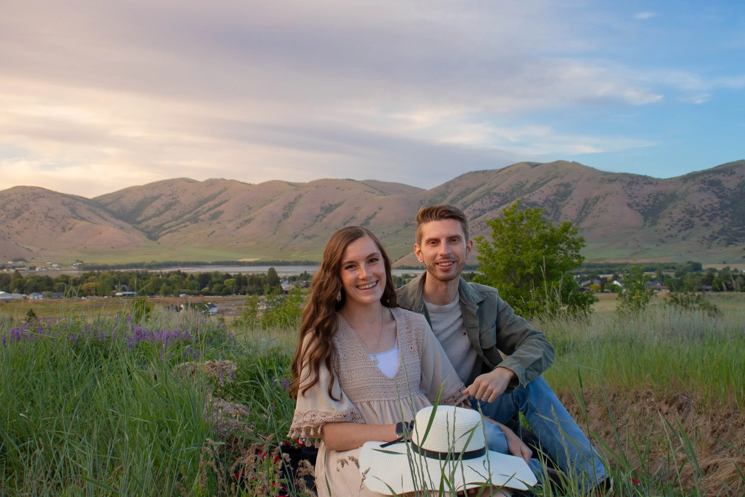 A smiling young woman and a young man sitting on grass in a field with mountains in the background during sunset.