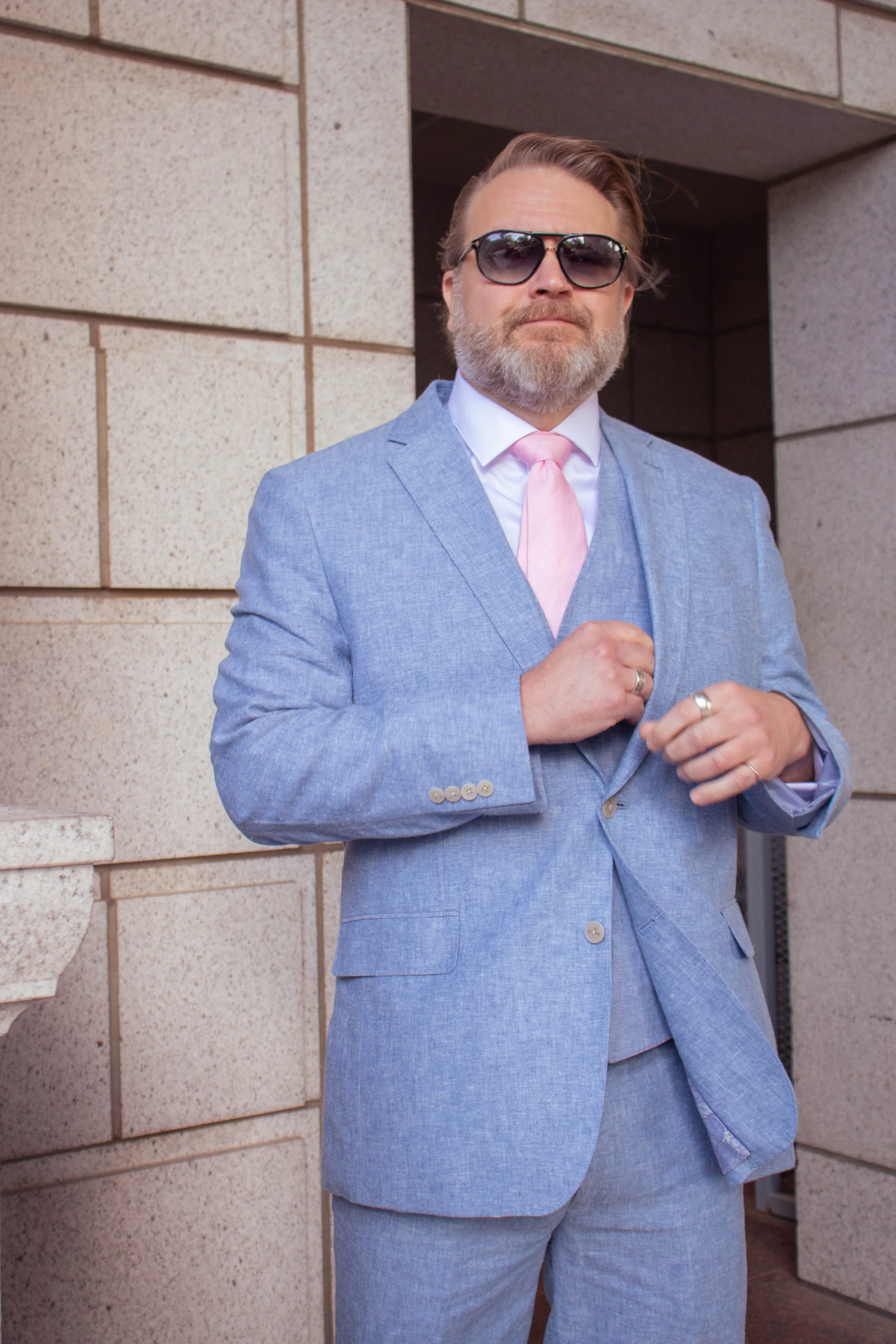 A man with light skin, gray beard, and sunglasses, dressed in a light blue suit with a pink tie, standing outdoors against a beige brick wall, adjusting his suit.
