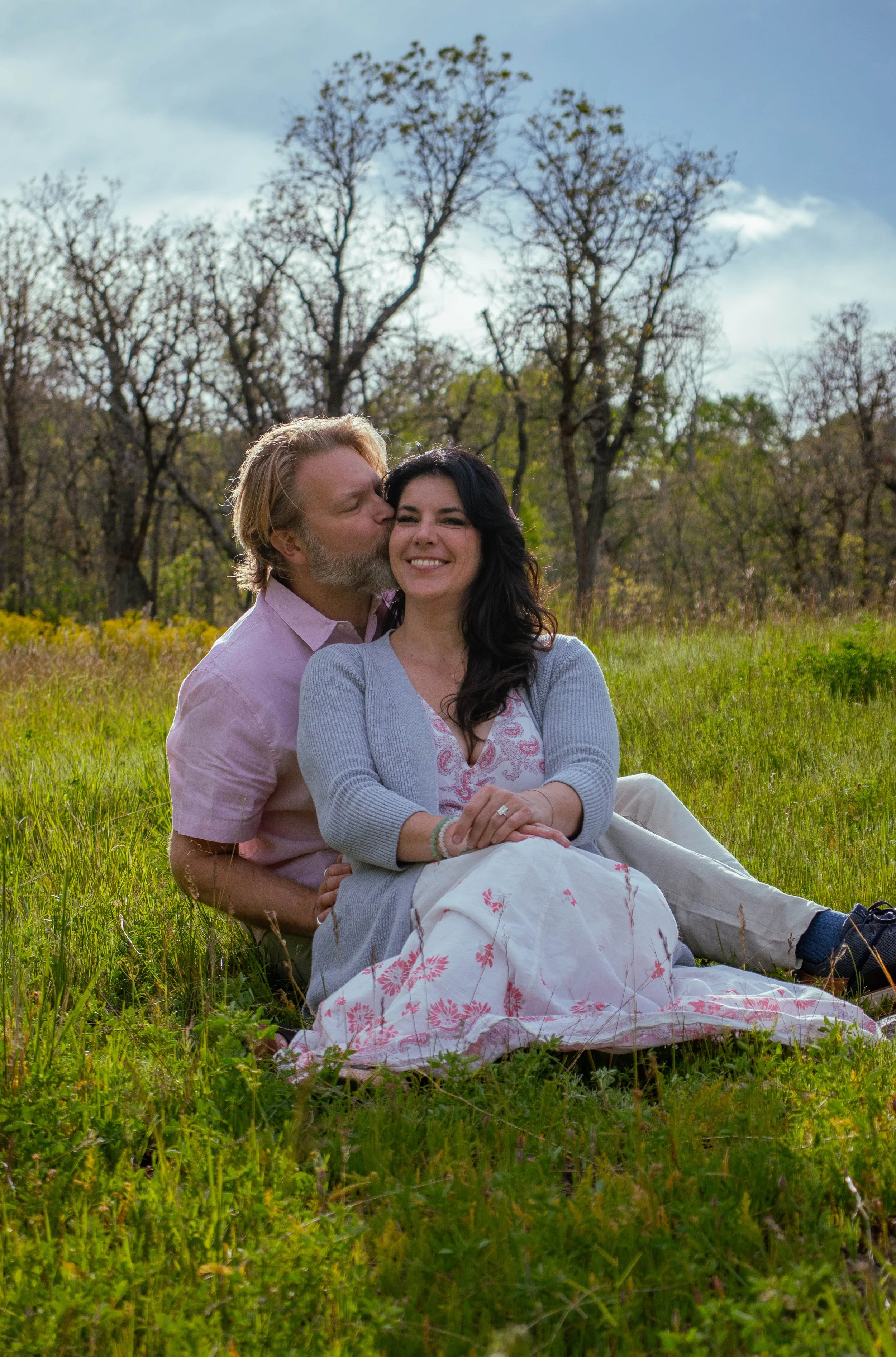 A man with a beard kissing a woman on the cheek while sitting together in a grassy field with trees in the background.