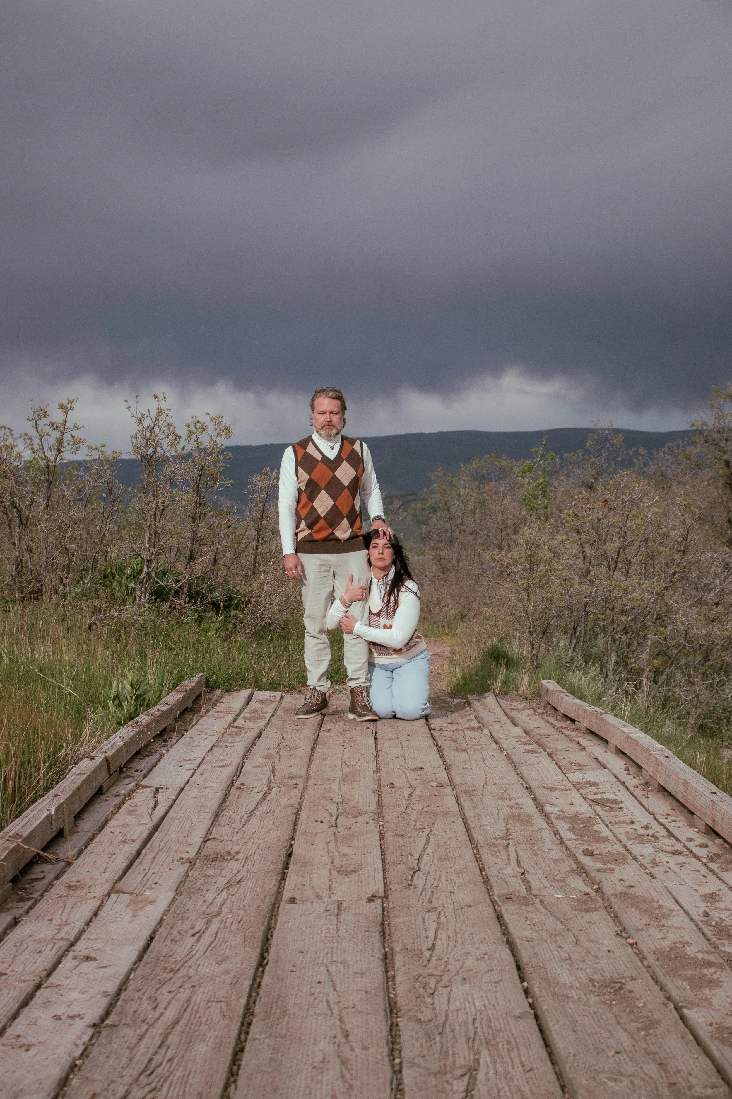 A man and a woman are on a wooden bridge in an outdoor setting with cloudy skies and mountains in the background. The man is standing with a serious expression, wearing a multicolored argyle sweater vest, white shirt, and beige pants. The woman is kn