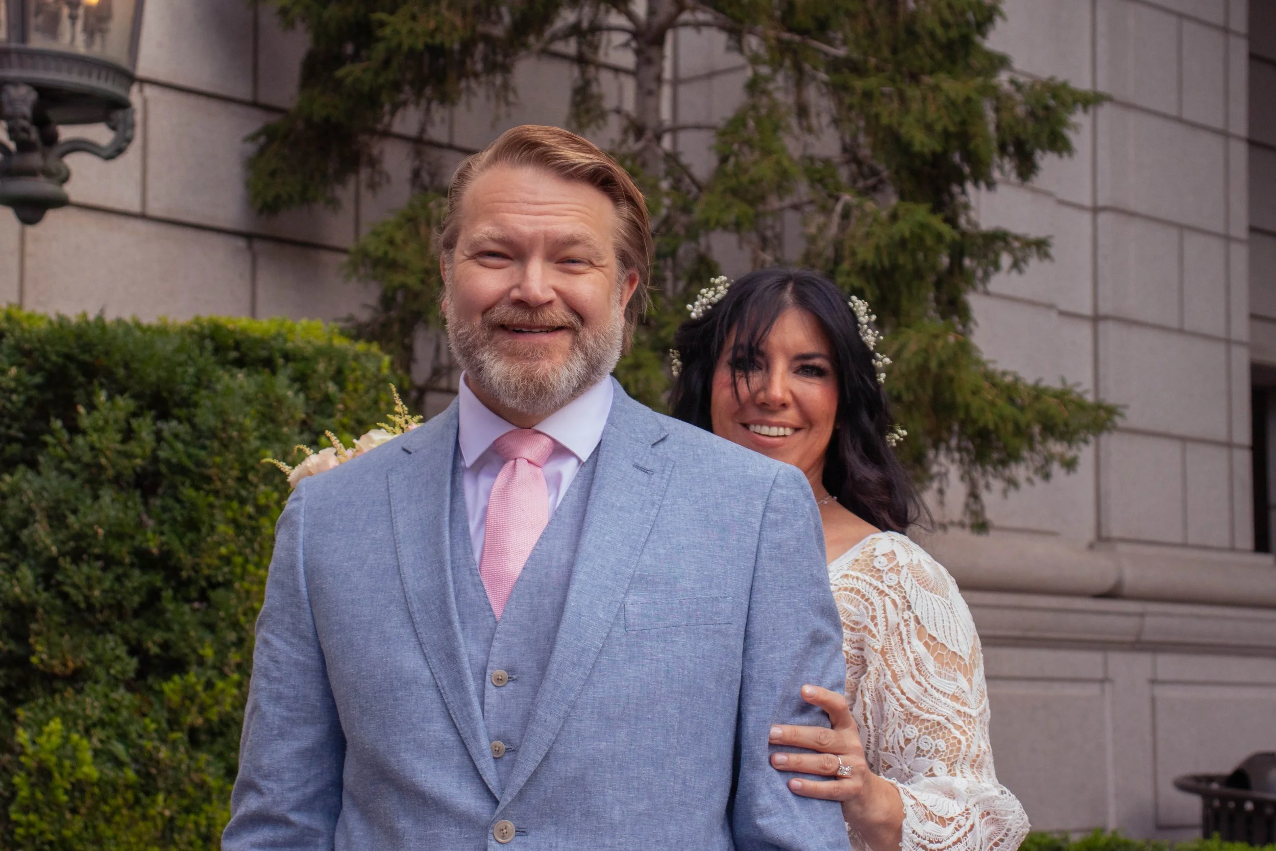 A smiling man in a light blue suit and pink tie is standing outside in front of a building with green shrubbery and a wall. Behind him, a woman with dark hair, wearing a white lace dress and small flower hair accessories, is hugging him from behind w