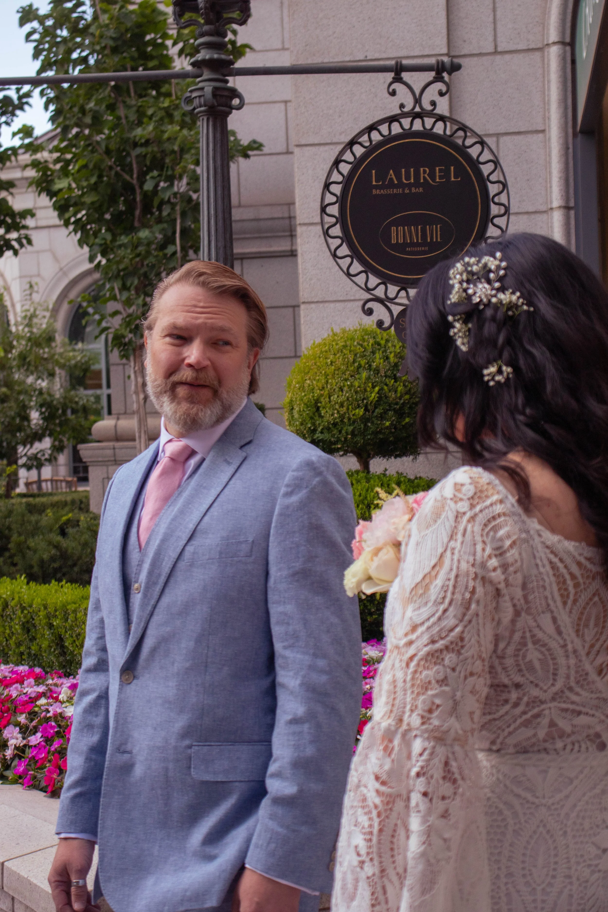 A man and a woman are standing outdoors near a building with a sign that reads 'Laurel Brasserie & Bar Bonne Vie Patisserie'. The man is wearing a light gray suit with a pink tie and has a beard and shoulder-length hair. The woman, whose face is not 