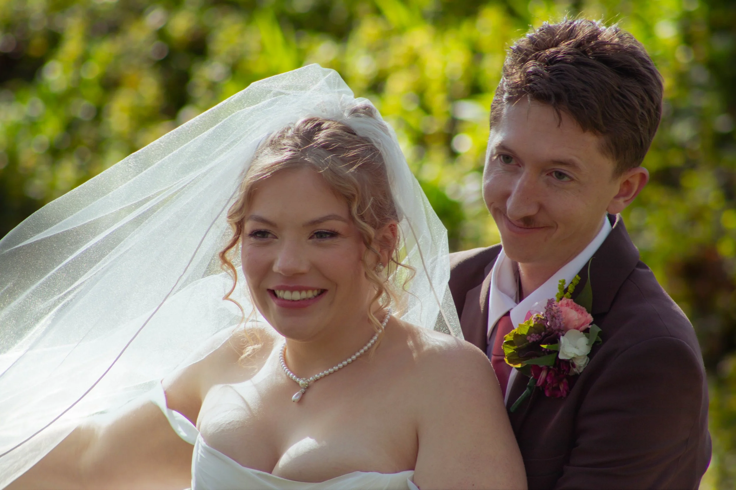 Bride and groom smiling, outdoors with green trees, bride in white dress and veil, groom in dark suit with boutonniere.