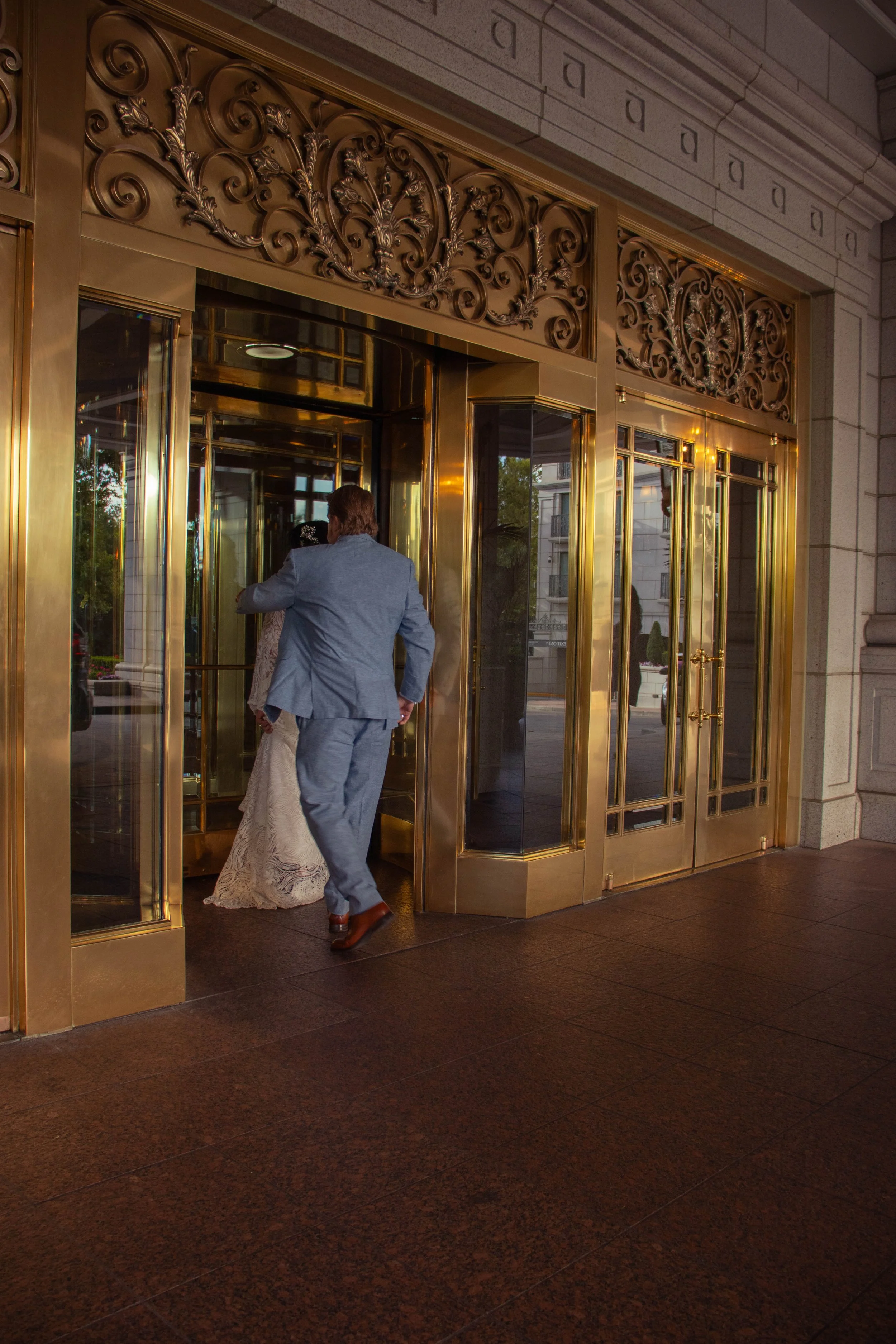 A man in a blue suit entering a building through an ornate gold and glass revolving door. A woman in a white lace dress is visible inside the building. The exterior features decorative gold detailing and stone architecture.